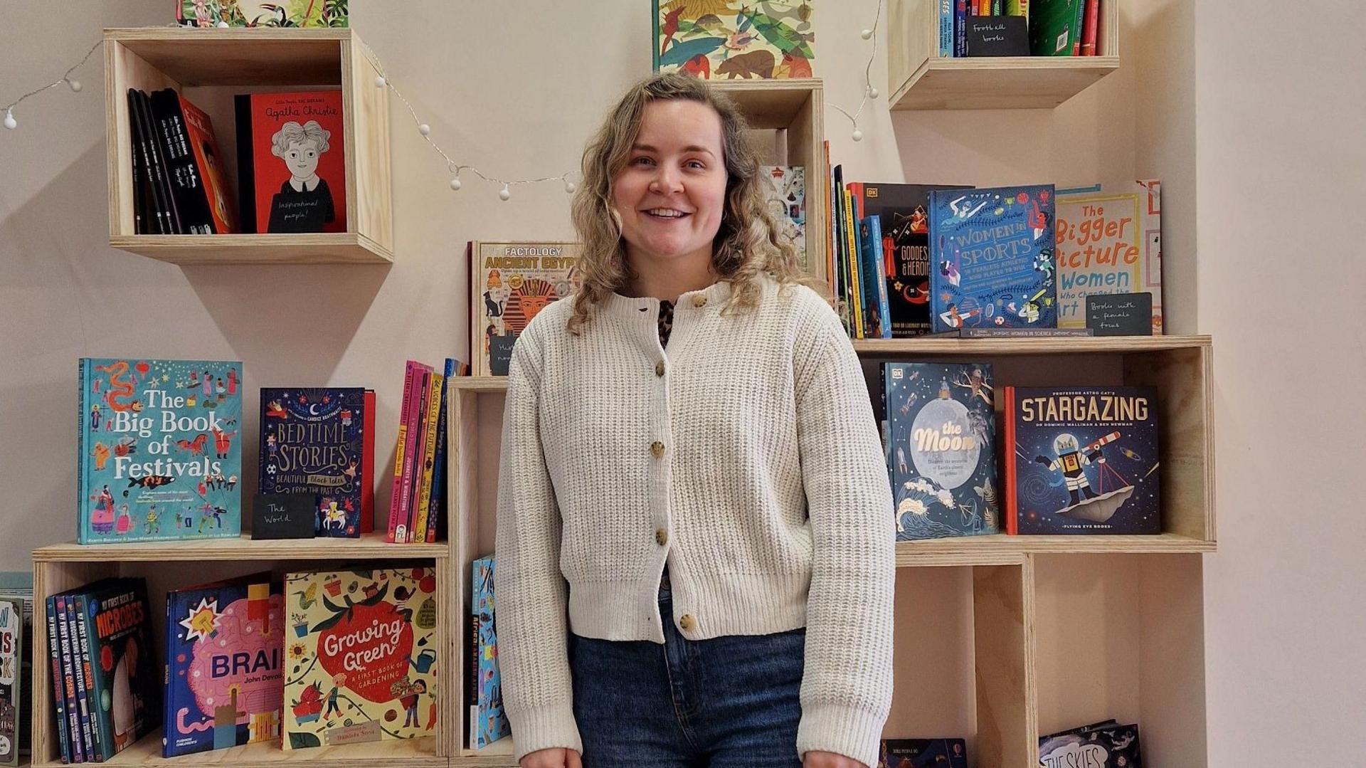 A woman with mid-length blonde curly hair wearing jeans and a white cardigan stands in front of a bookshelf.