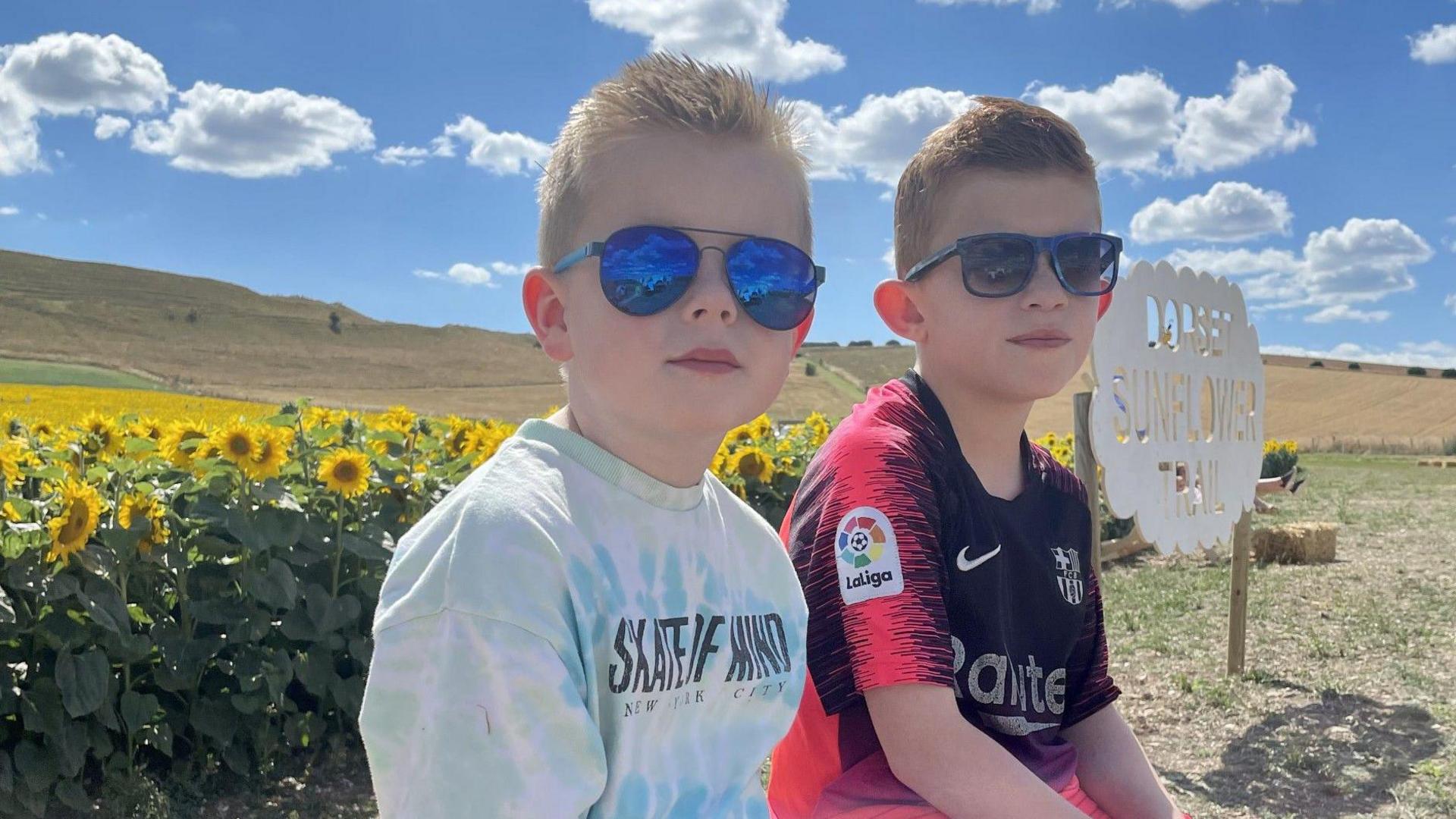 (l-r) Leo and Archie, two brothers sitting on a hay bail with sunflowers in the background. Both boys are wearing t-shirts and sunglasses.