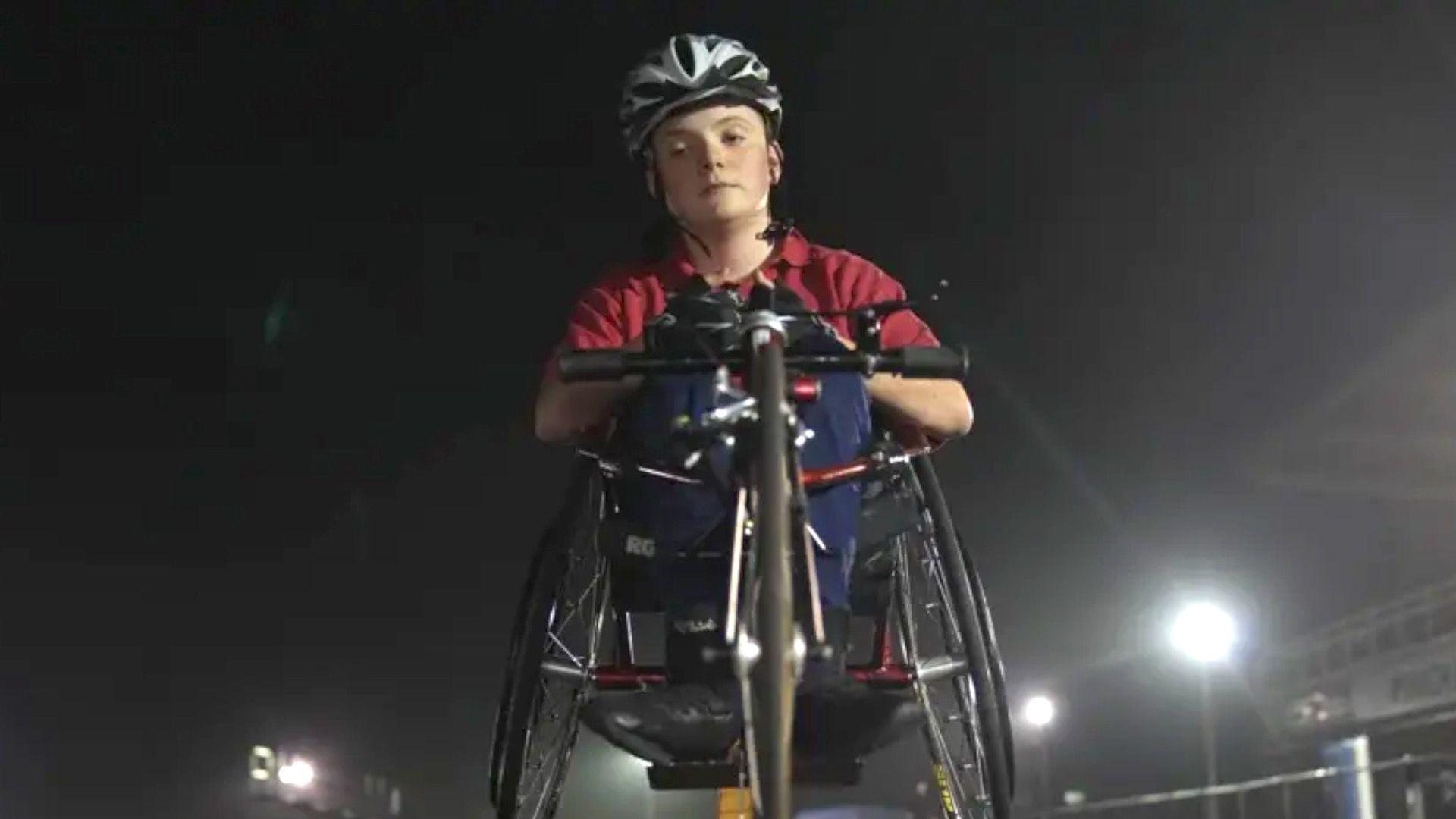 Morgan is pictured in his racing wheelchair on the athletics track. He is looking down at the camera with a determined expression on his face. He is wearing a silver helmet and a red t-shirt with racing gloves on his hand.