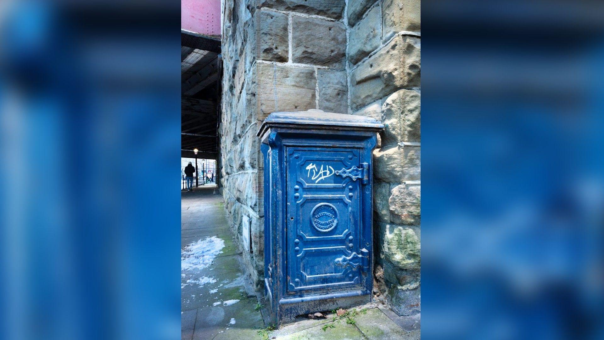 A blue electricity box stands alongside a traditional stone wall. It is tagged with white graffiti.