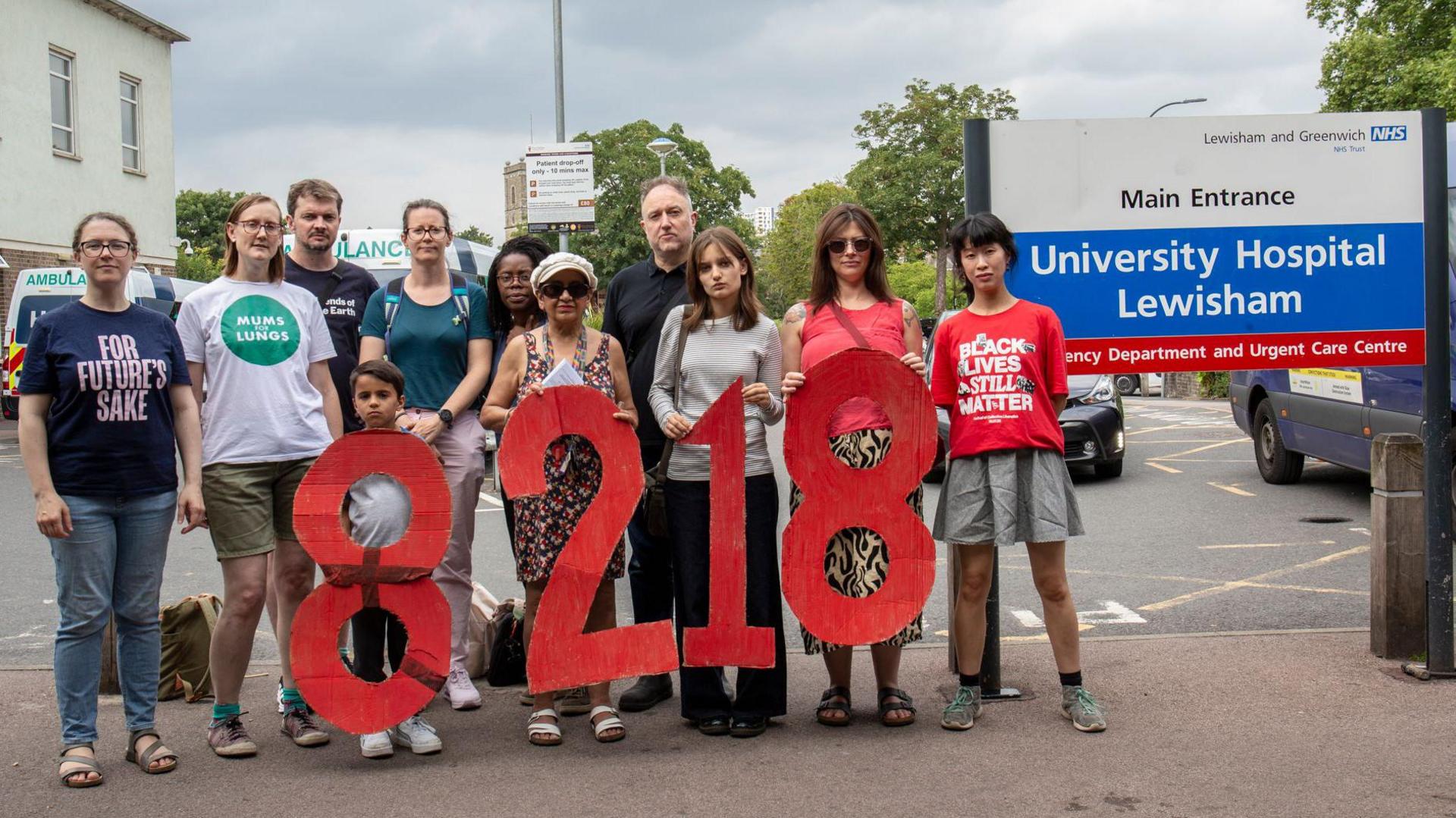 A group of men and women, and one child, some wearing Mums for Lungs tops, are stood holding large numbers 8218, in red. They are stood by the entrance of University Hospital Lewisham.