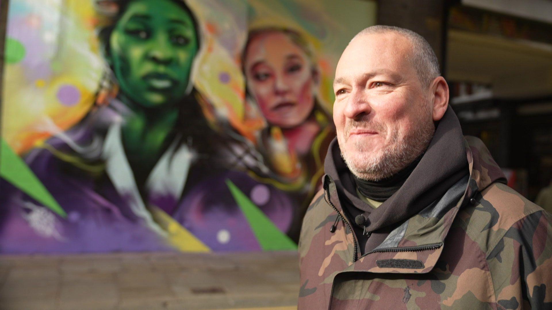Mr Cenz stands in front of his mural, showing the main characters from Wicked