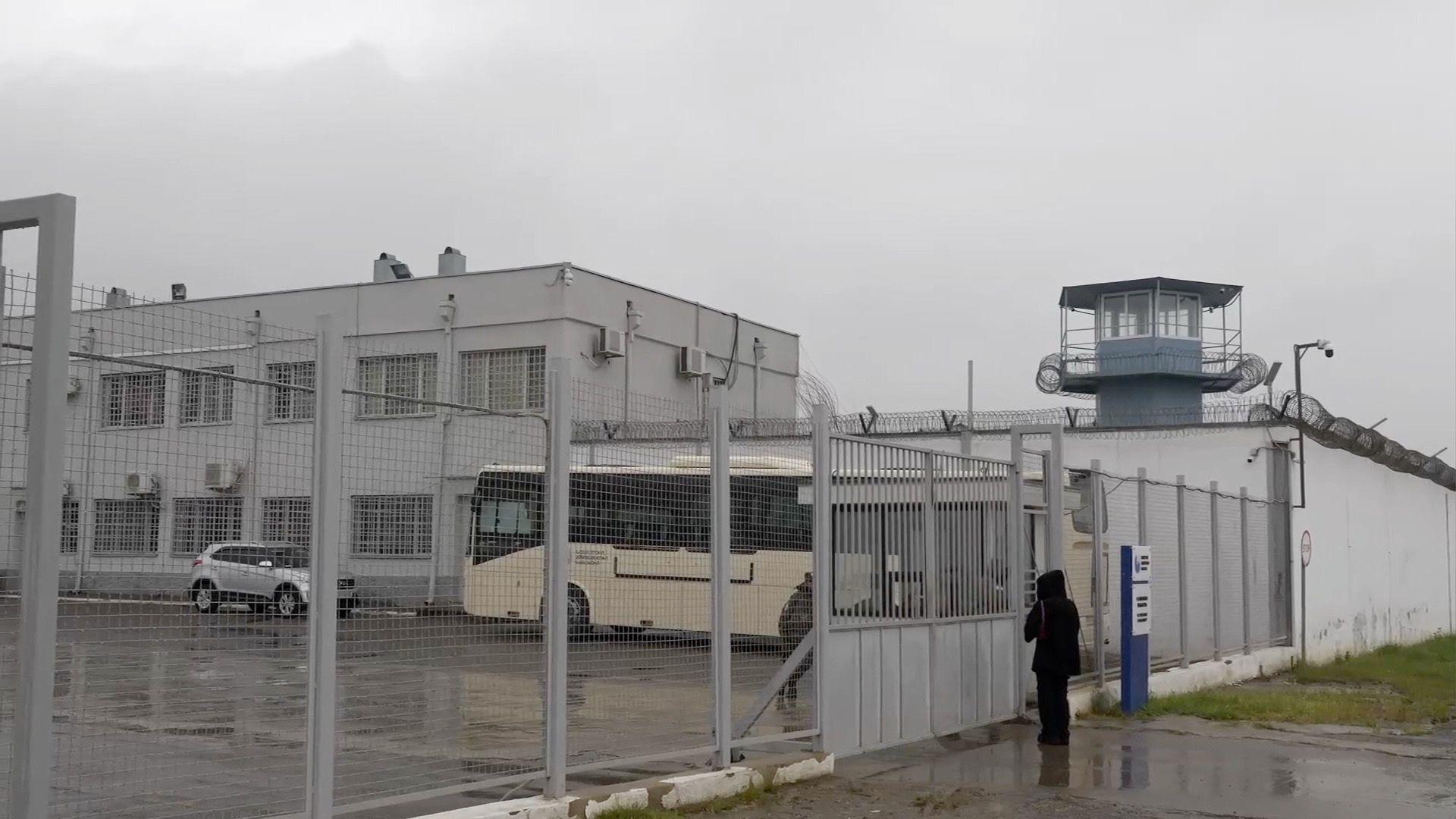 An outside view of the prison. A coach and a car are waiting just inside the gates. A figure wearing black can be seen standing just outside the gates. The prison has lots of barbed wire.