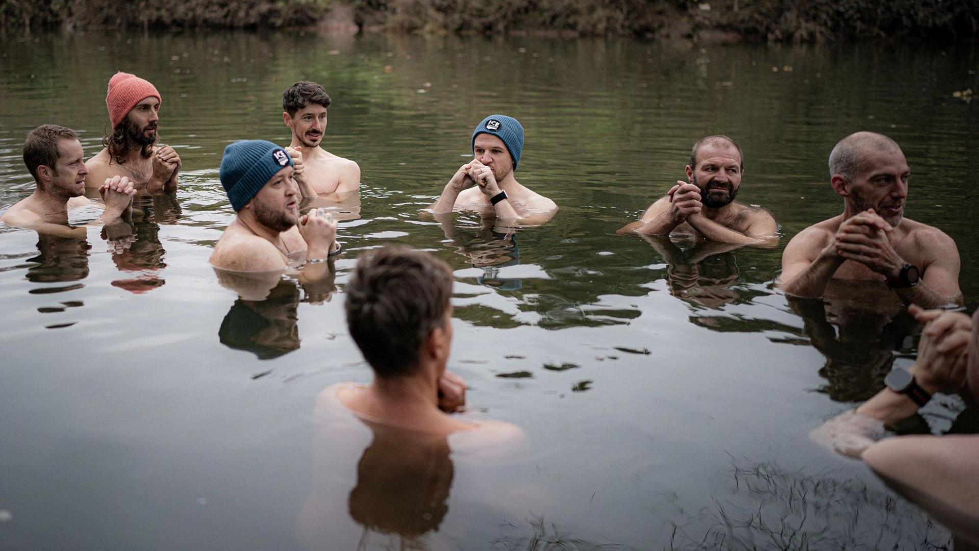 A group of men, some wearing woolly hats, stand in the River Avon at Conham in Bristol. It is an autumn day and they are all clasping their hands near their chins. The water is dark green and fairly still.