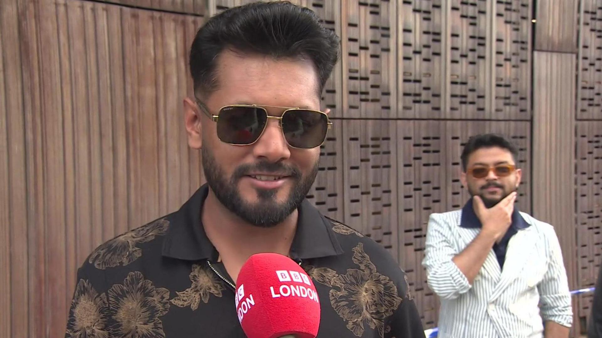 A man in dark glasses and a dark shirt with a brown floral design is smiling. A red microphone with the words BBC London is in front of him.
Another young man in a white suit with stripes and a black shirt can be seen to the right.