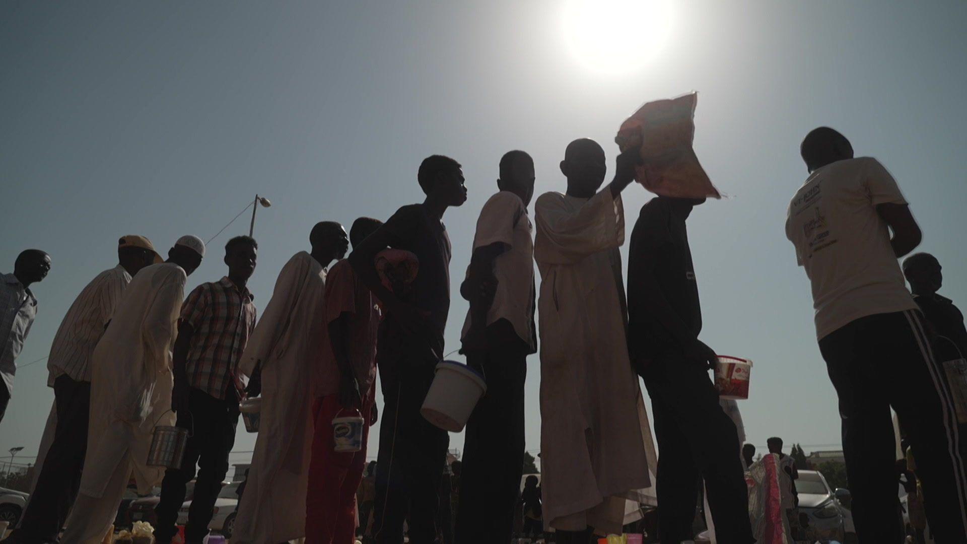 The silhouettes of a group of people holding buckets and waiting in a queue.