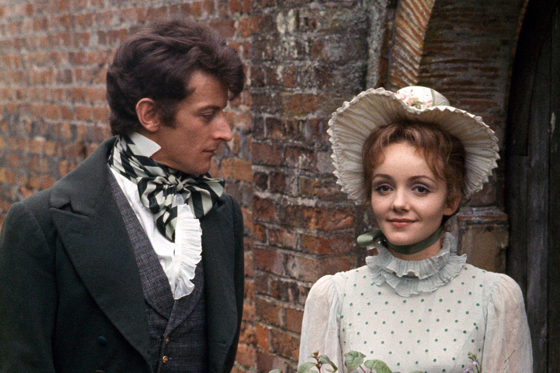 a still featuring lewis fiander as mr darcy in a black coat, grey waistcoat and white frilly shirt with a striped grey cravat. he looks at celia bannerman as elizabeth bennet in a white high necked dress with green polka dots and a matching white bonnet