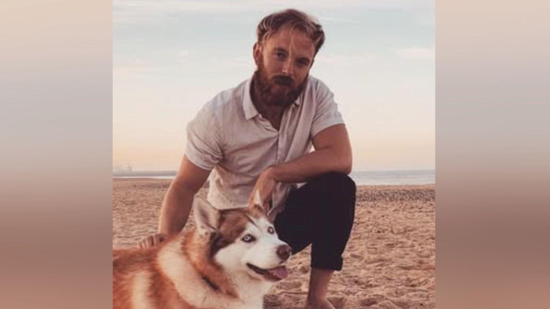 Benjamin Gladman kneeling on a beach with a dog. Mr Gladman, who has light brown hair and a beard, is wearing a beige short-sleeved shirt and blue trousers. He is barefoot. He has his right hand on the back of the dog, which is a German Shepherd-type breed.