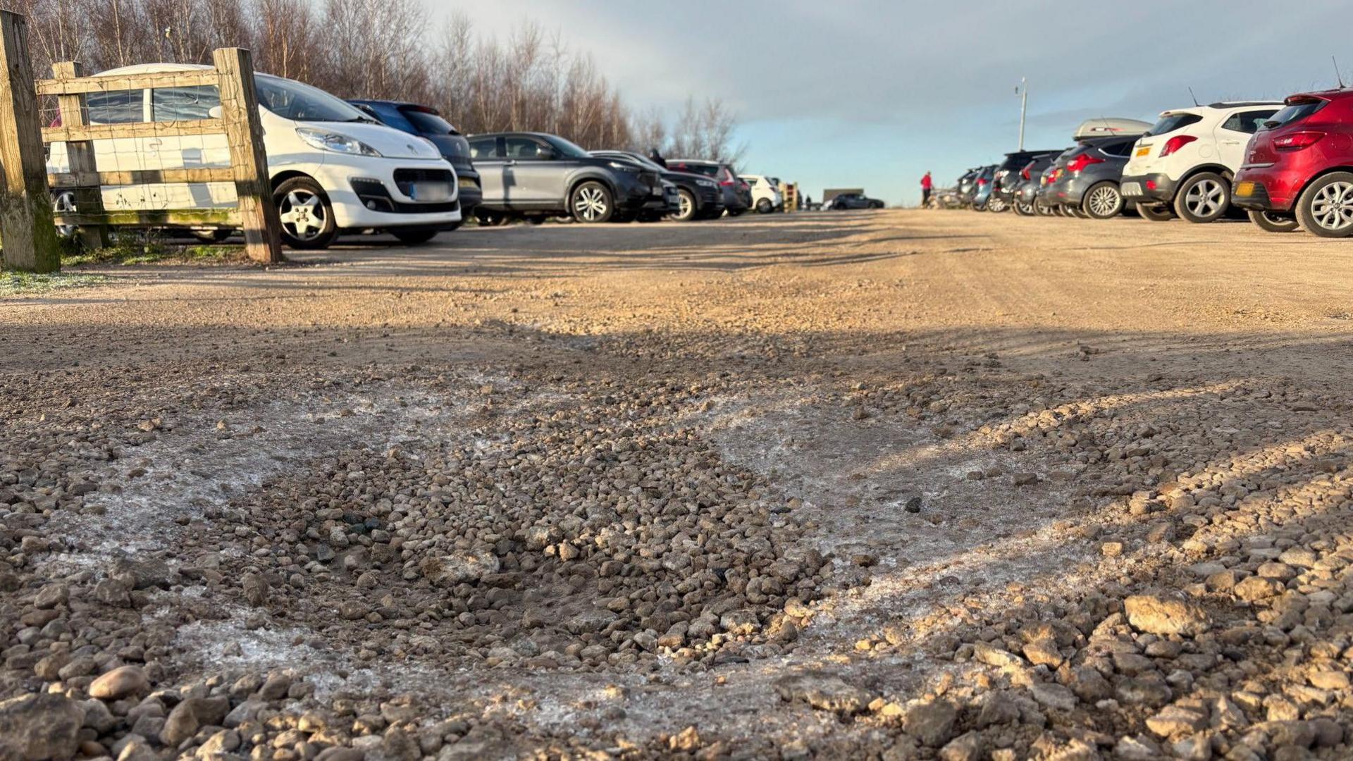 Ground level shot of Gedling Country Park car park, with a pothole in the foreground and about 20 cars parked on either side of a gravel road