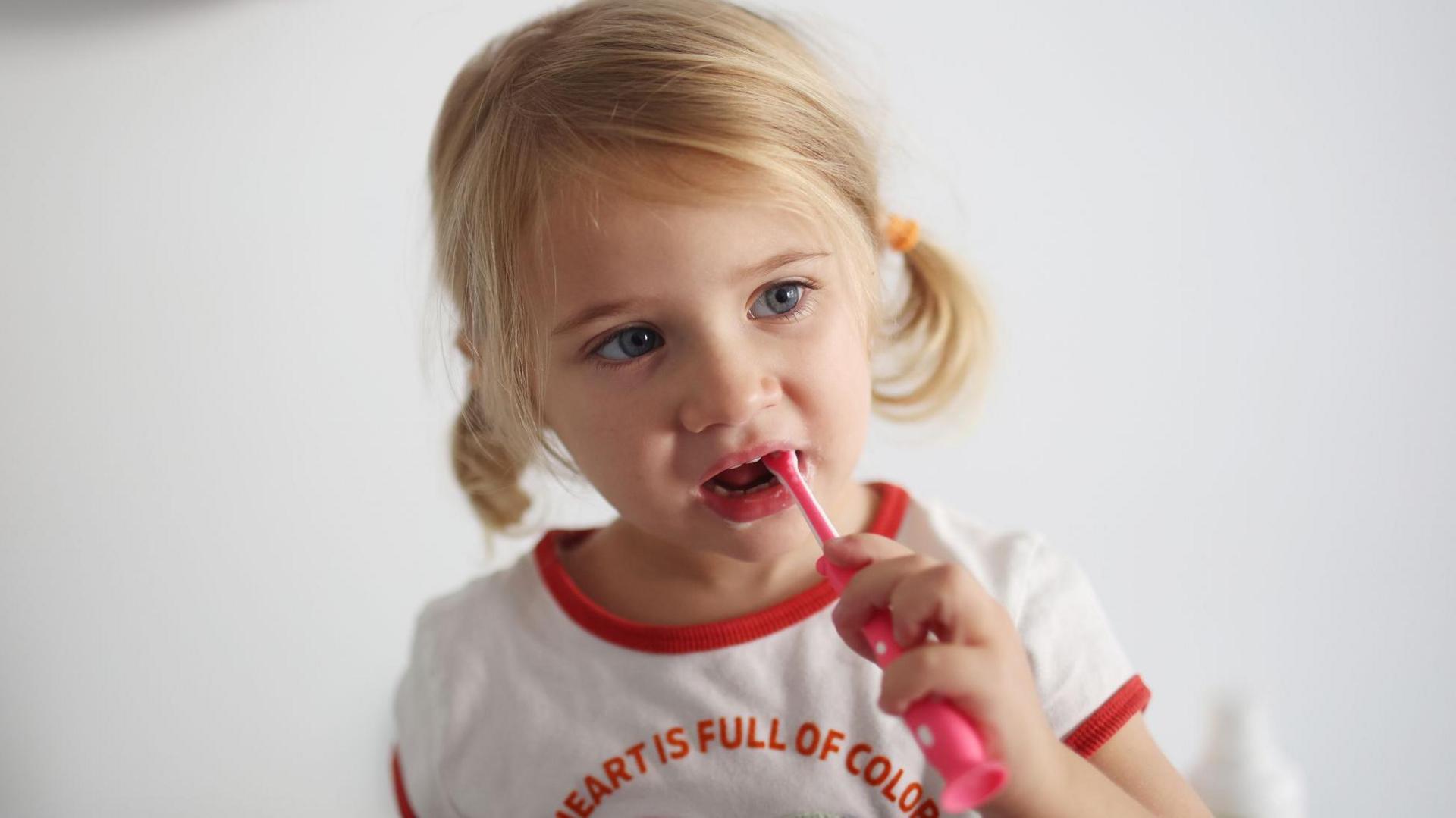 A young girl with blonde hair tied back in pigtails brushes her teeth. She is wearing a white t-shirt which says My heart is full of colours