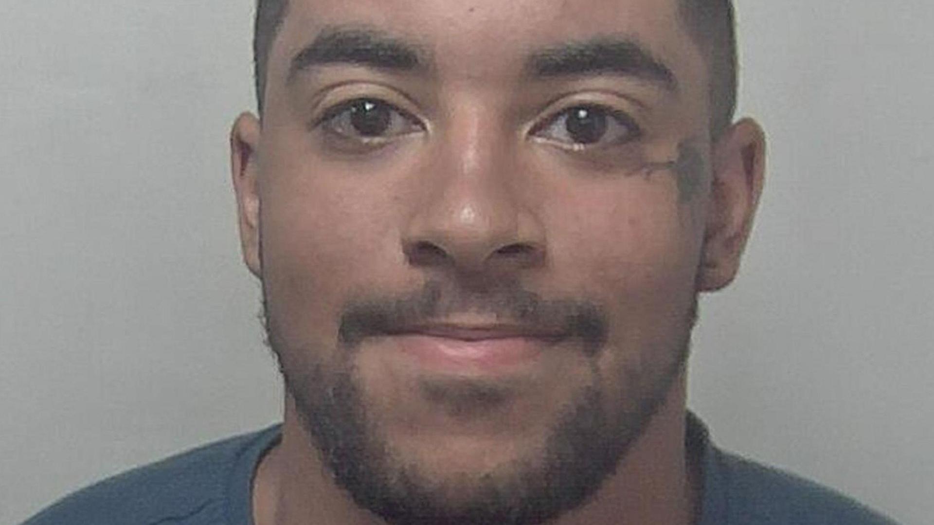 A custody photo of a man with short brown hair and facial hair, and brown eyes. He has a small tattoo to the left of his left eye and is wearing a dark blue t-shirt