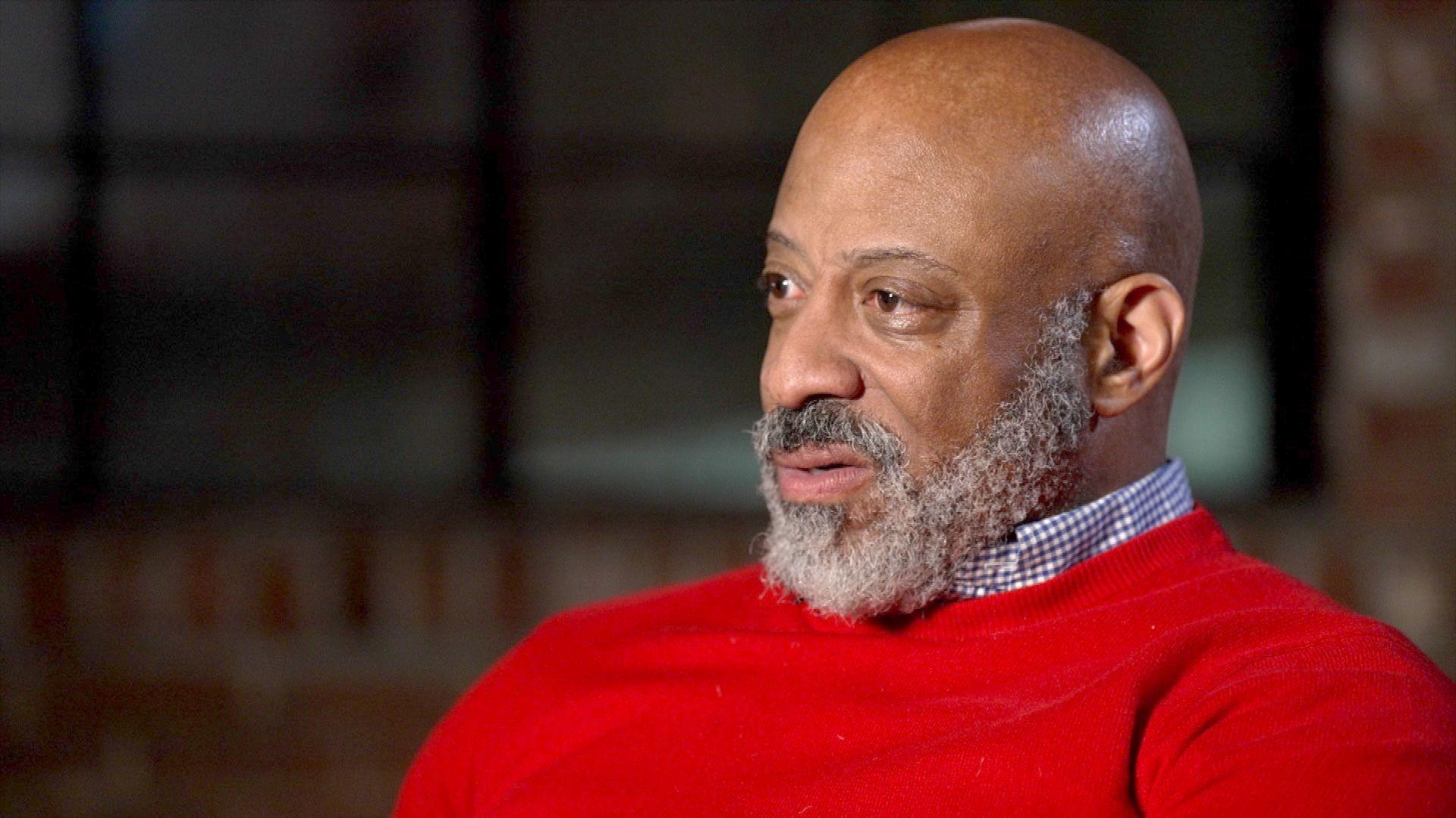 Daniel Evans, a man with a shaved head and a graying beard, who wears a bright red jumper and a blue-and-white checked shirt, spotlit in a room with bare brick walls and large warehouse-style windows just visible in the darkened background