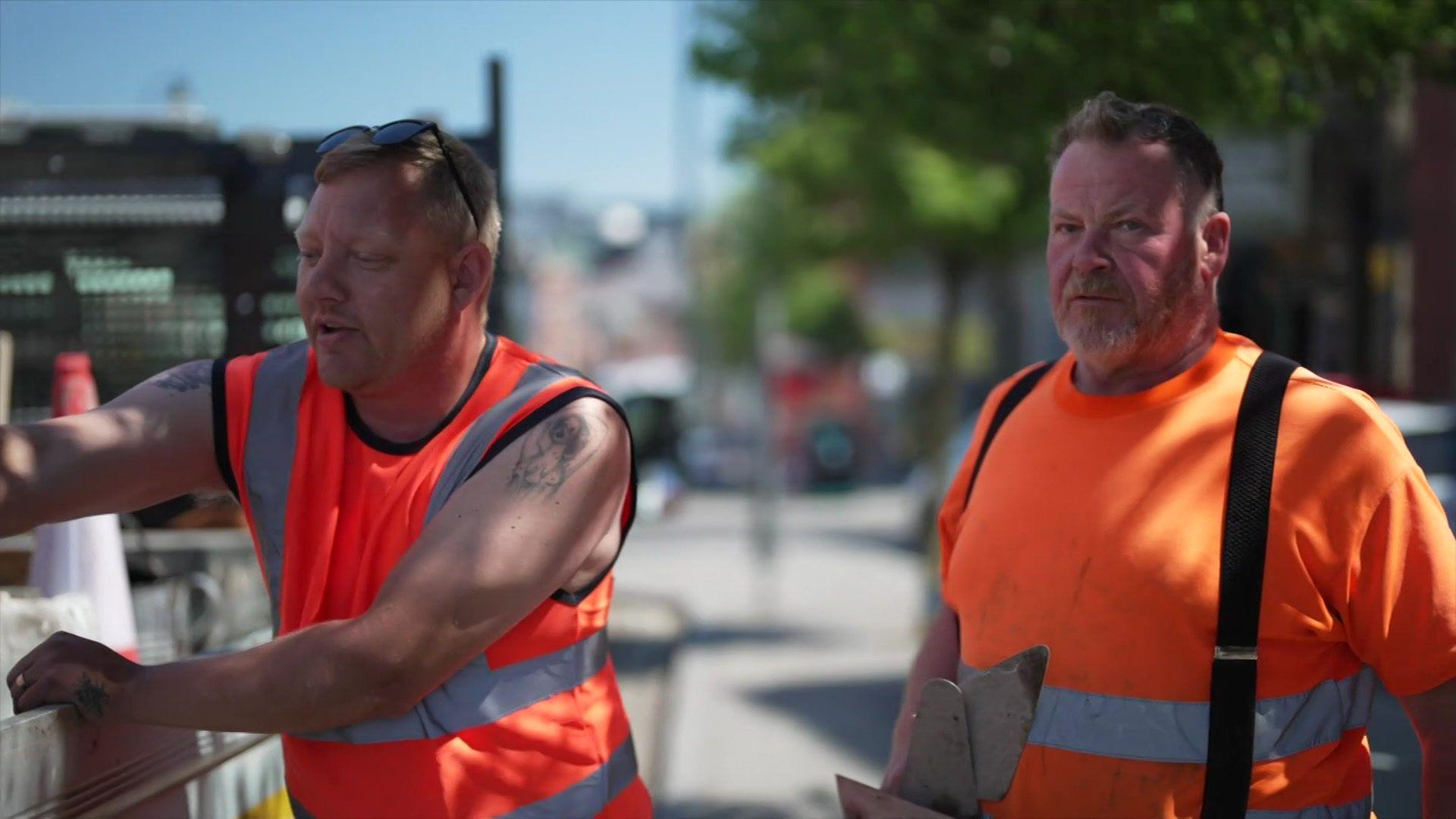 Two men wear high-vis orange clothes as they work by the side of the road
