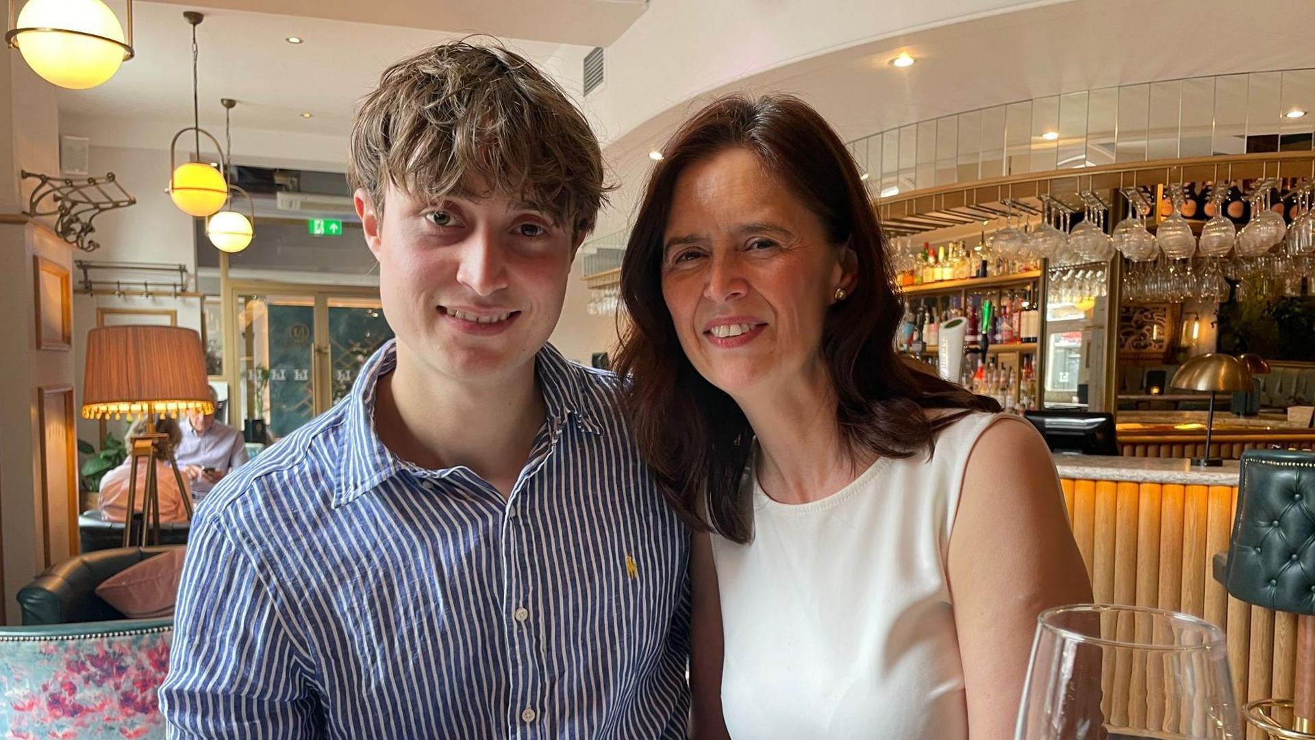 A man and his mother look at the camera whilst smiling. They are sat in a modern bar.