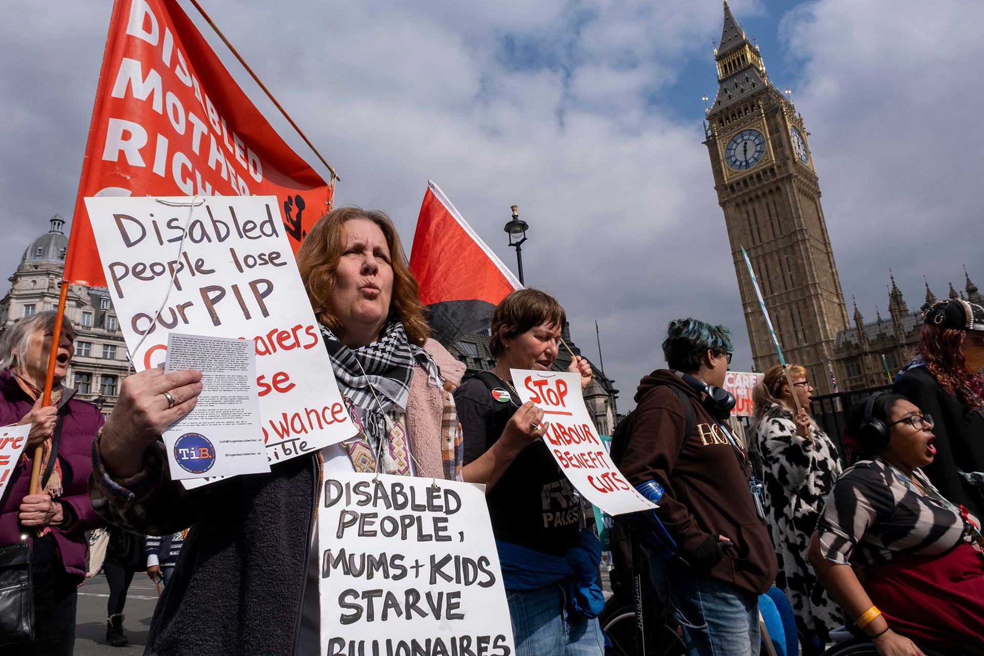 A group of protesters hold signs, with two of the most legible reading 'disabled people lose our PIP' and 'disabled people, mums + kids starve', as they walk along Parliament Square with Big Ben visible in the background against a cloudy blue sky on 26 March.