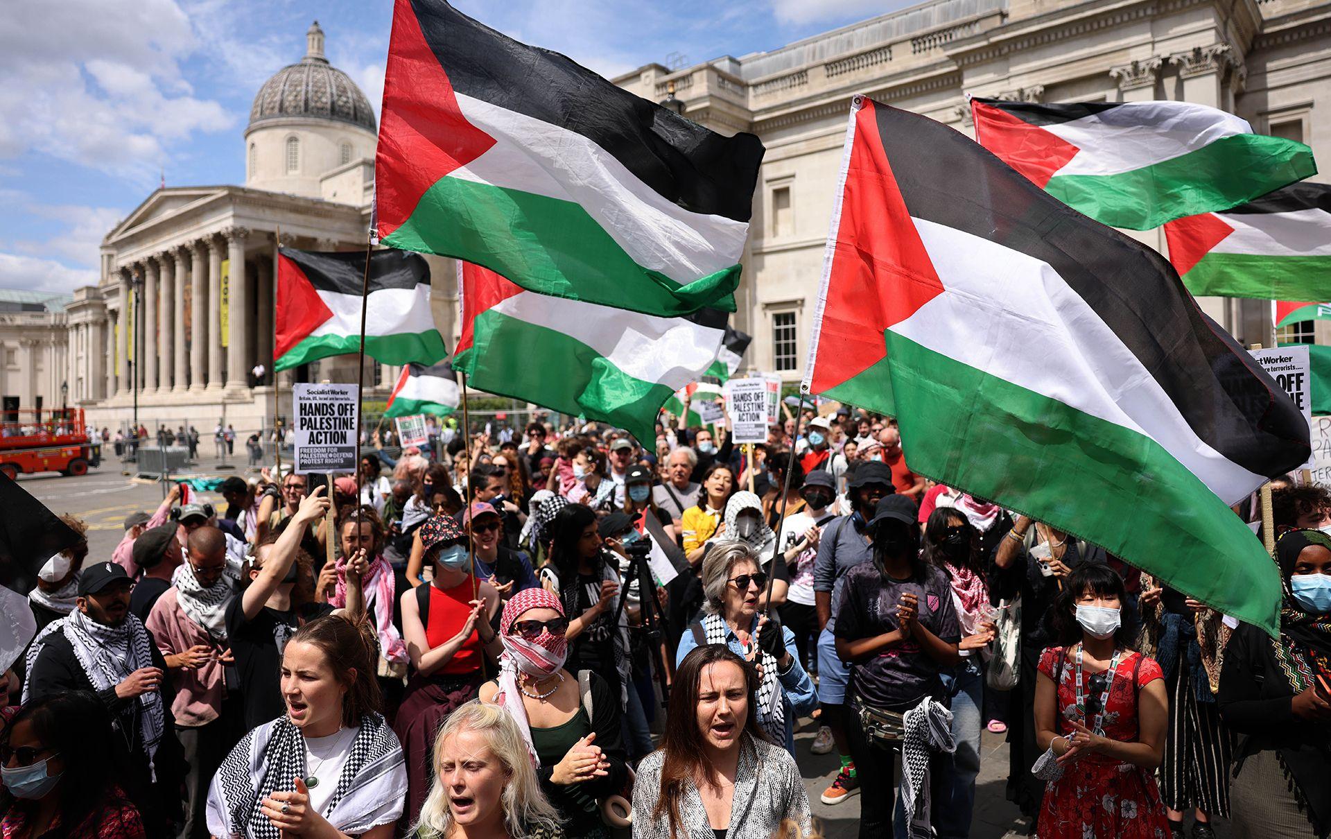 A crowd of people, with many holding Palestinian flags, as they look forward behind the camera. It looks like a lot of them are shouting, and the National Gallery can be seen in the background against a blue sky.