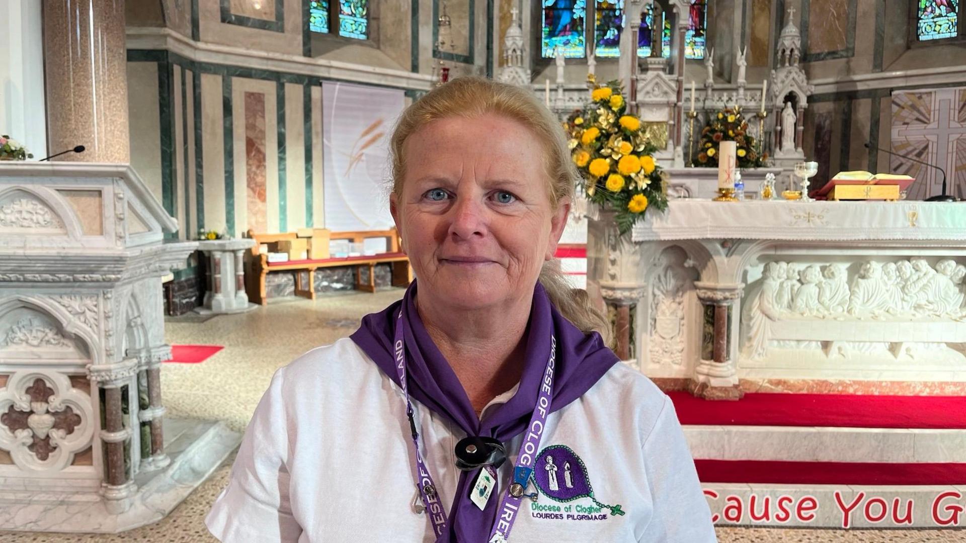 Olga Gilliland has blonde hair pulled back into a ponytail. She is wearing a white T-shirt and a purple scarf. She is standing in front of the altar of the church