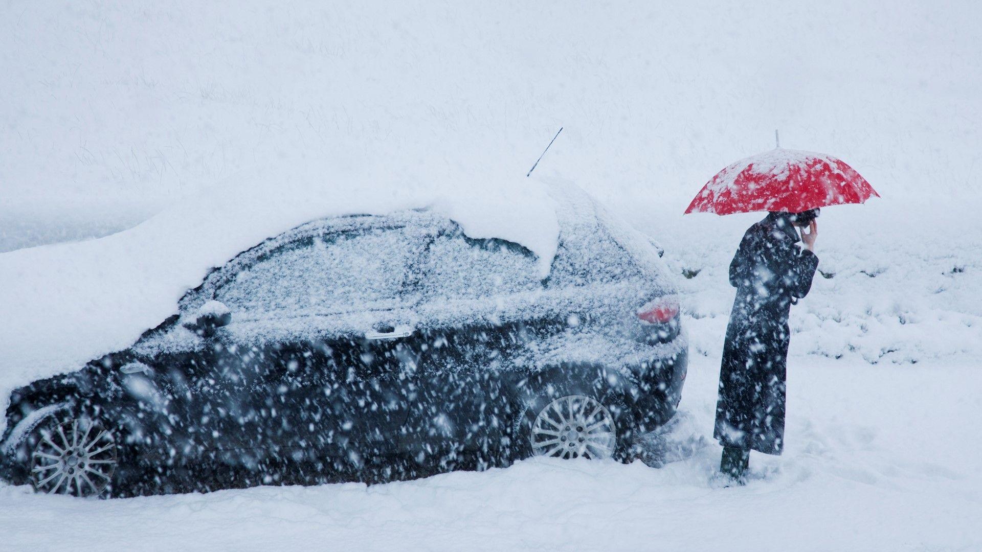 A person in a long winter coat holding a red umbrella beneath falling snow stands next to a snow-covered car with thick snow lying on the ground.  