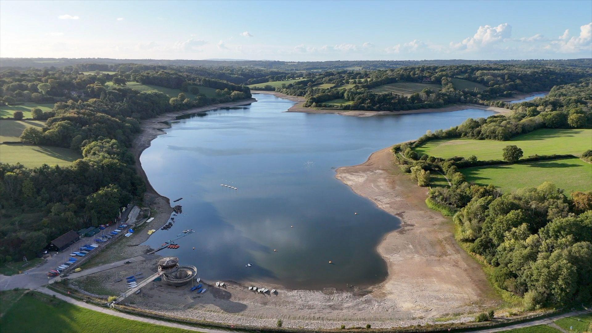 An aerial shot shows a reservoir surrounded by green fields and trees. You can see large areas of brown around the edges of the reservoir where the water has receded.
