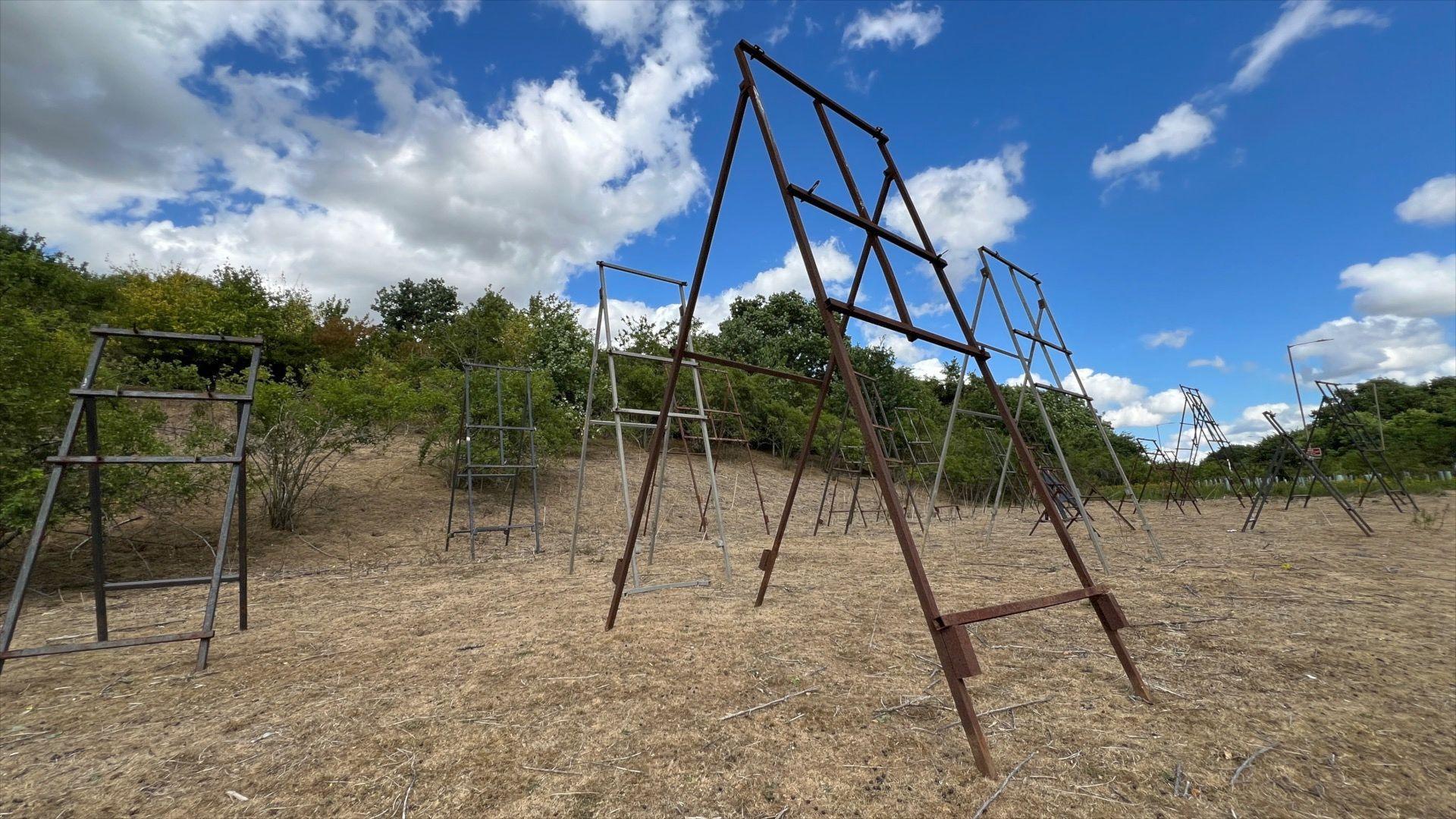 Several metal A-frames standing upright, all facing the same direction. They each have four legs but do not have any road signs displayed on them. They are standing on a patch of grass with trees and bushes behind them and blue skies above