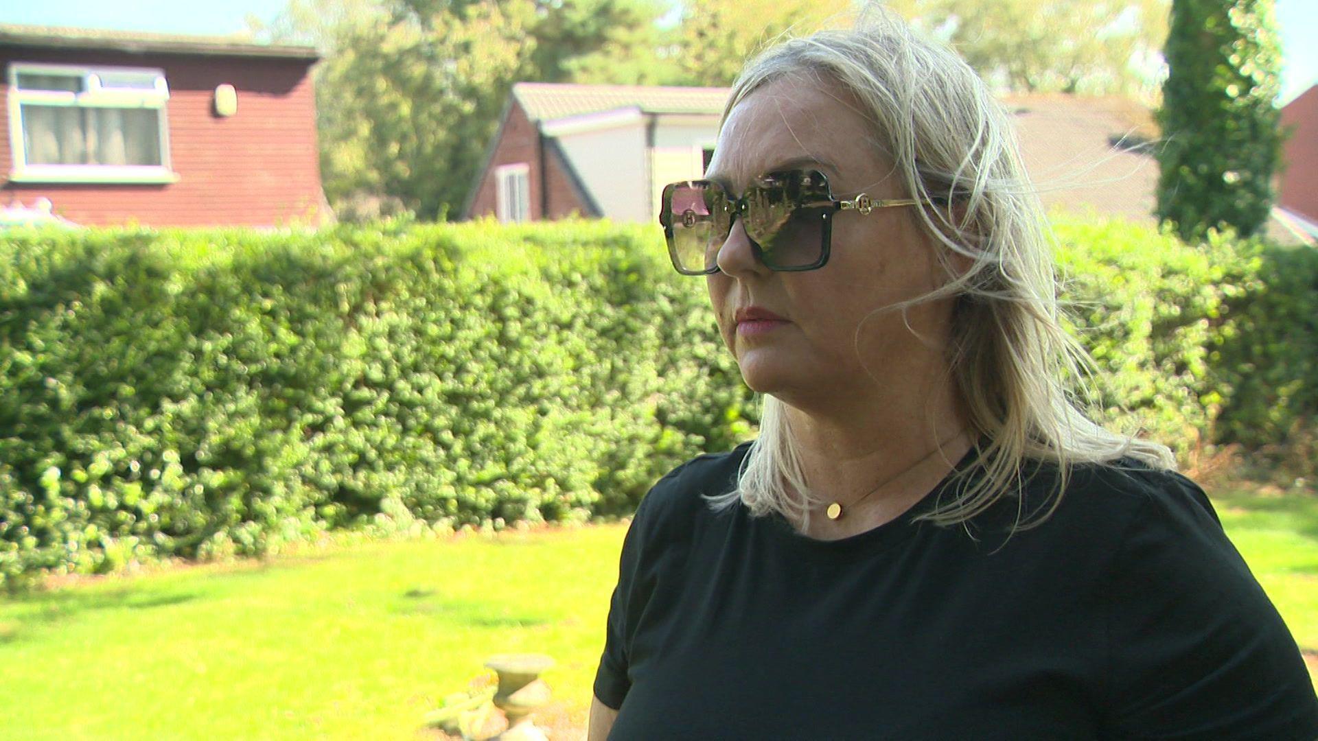 Joanne has blonde hair and wears black, and has sunglasses on as she is pictured next to the mass grave where her uncle is buried. 