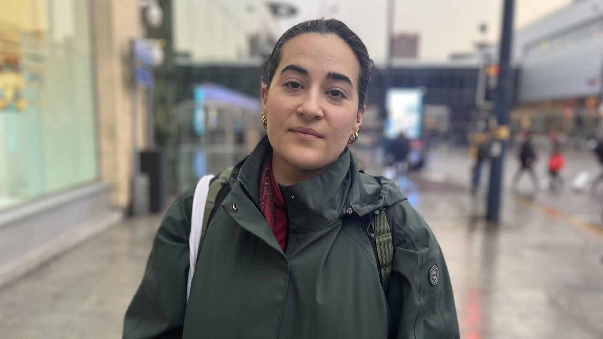 A woman with dark tied back hair and  wearing gold earrings and a dark green waterproof coat is standing on a busy street blurred in the background.