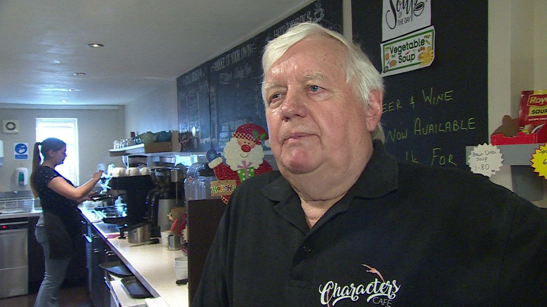 A man wearing a black polo shirt standing in a cafe, with a blackboard menu and a member of staff using a coffee machine behind him.