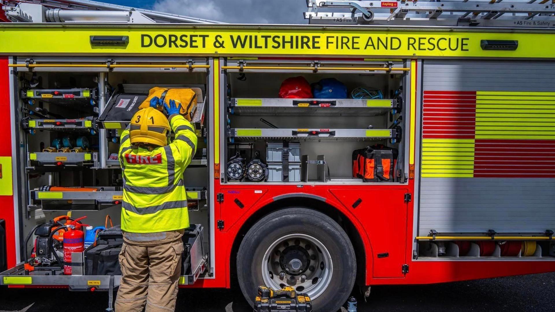 A firefighter wearing fireproof brown trousers, a hi-vis yellow jacket and a yellow helmet. They are returning kit back into the side of a parked fire engine, which has it's shutters pulled up. 