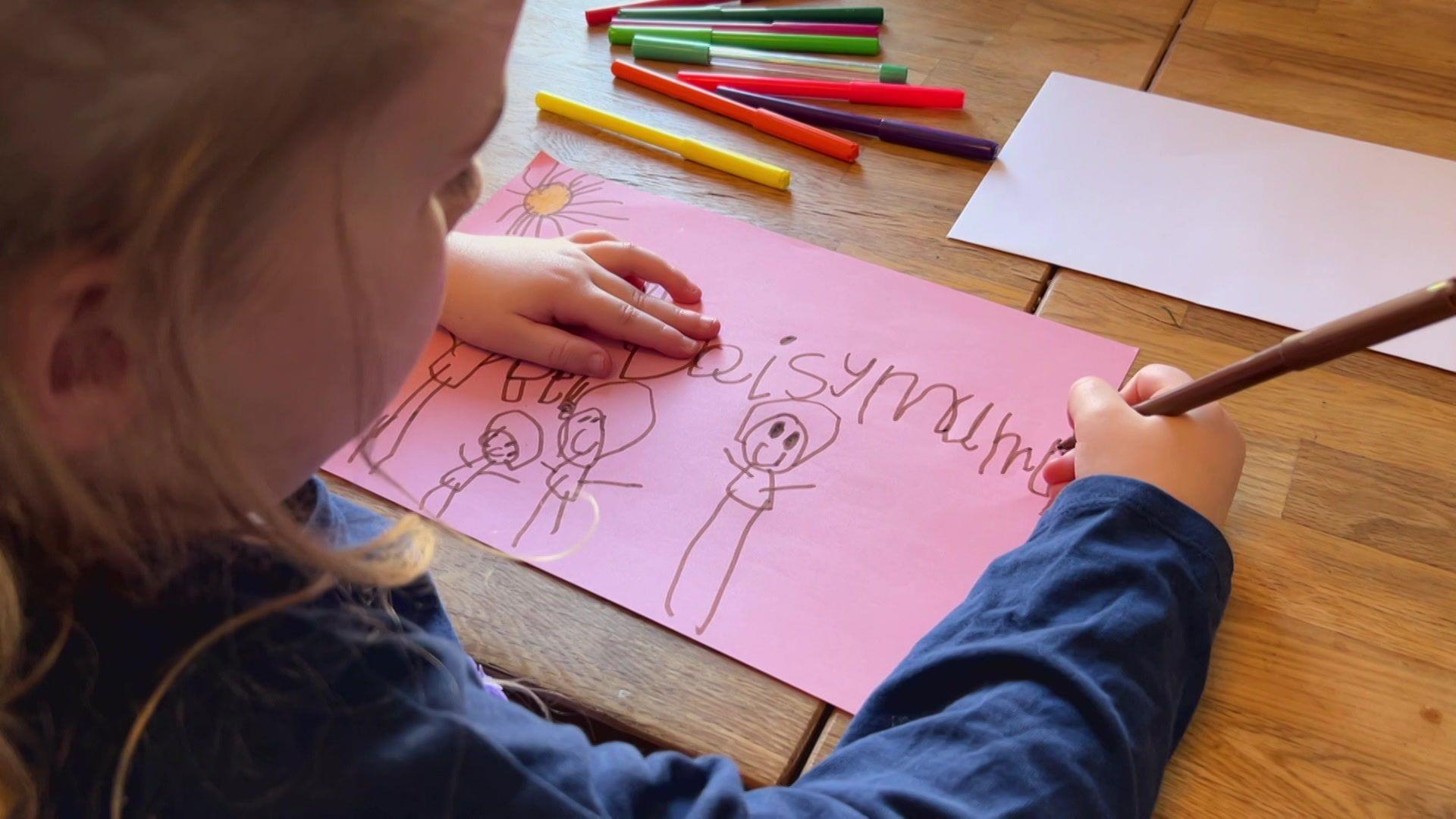 Daisy drawing a picture for her nan with coloured felt-tip pens, the picture is of all the family with the word "mummy".