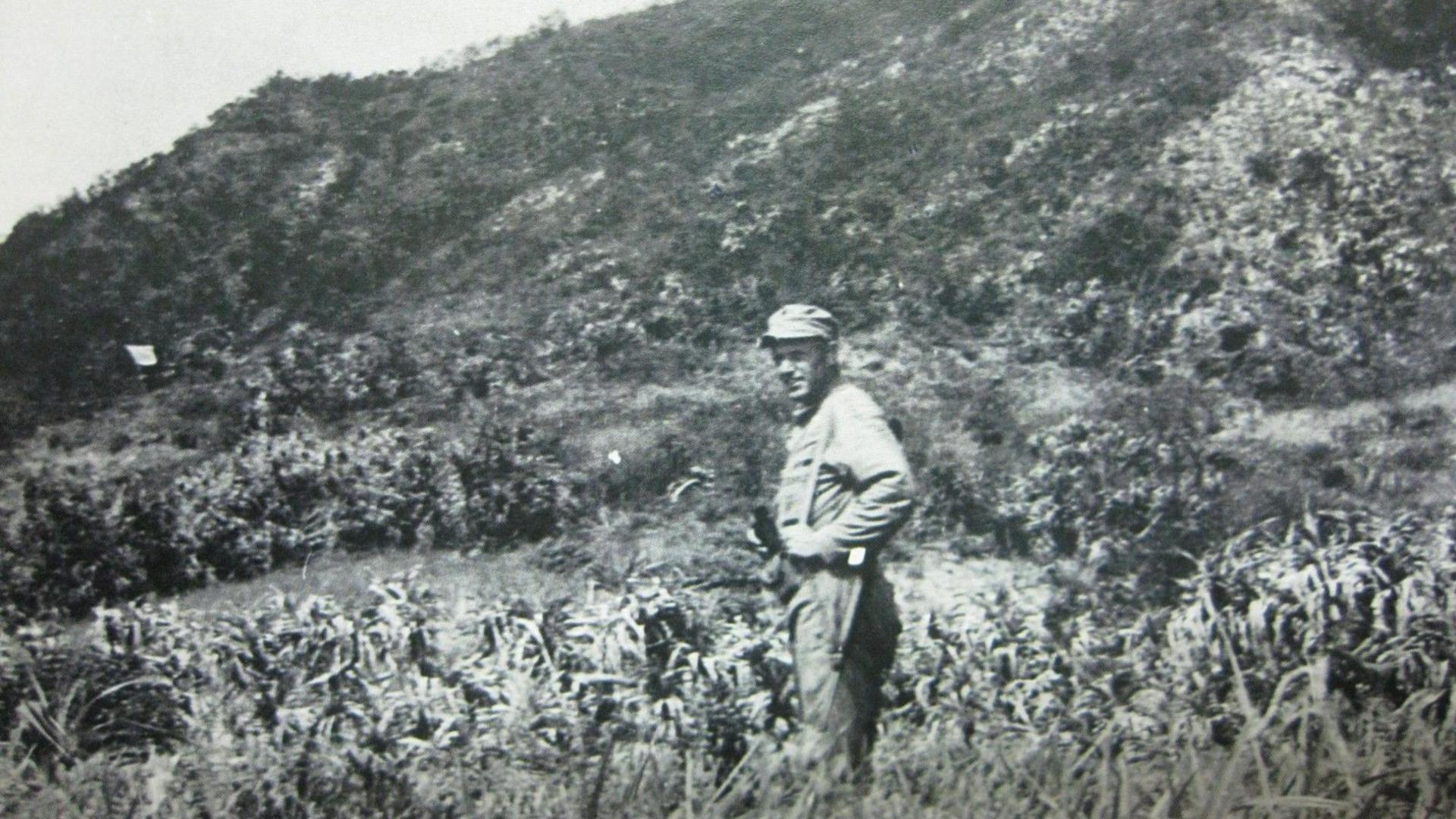 A black and white image of George Clark standing on Aka island in front of a cliff. To one side, a row of troops carrying a white flag is just visible