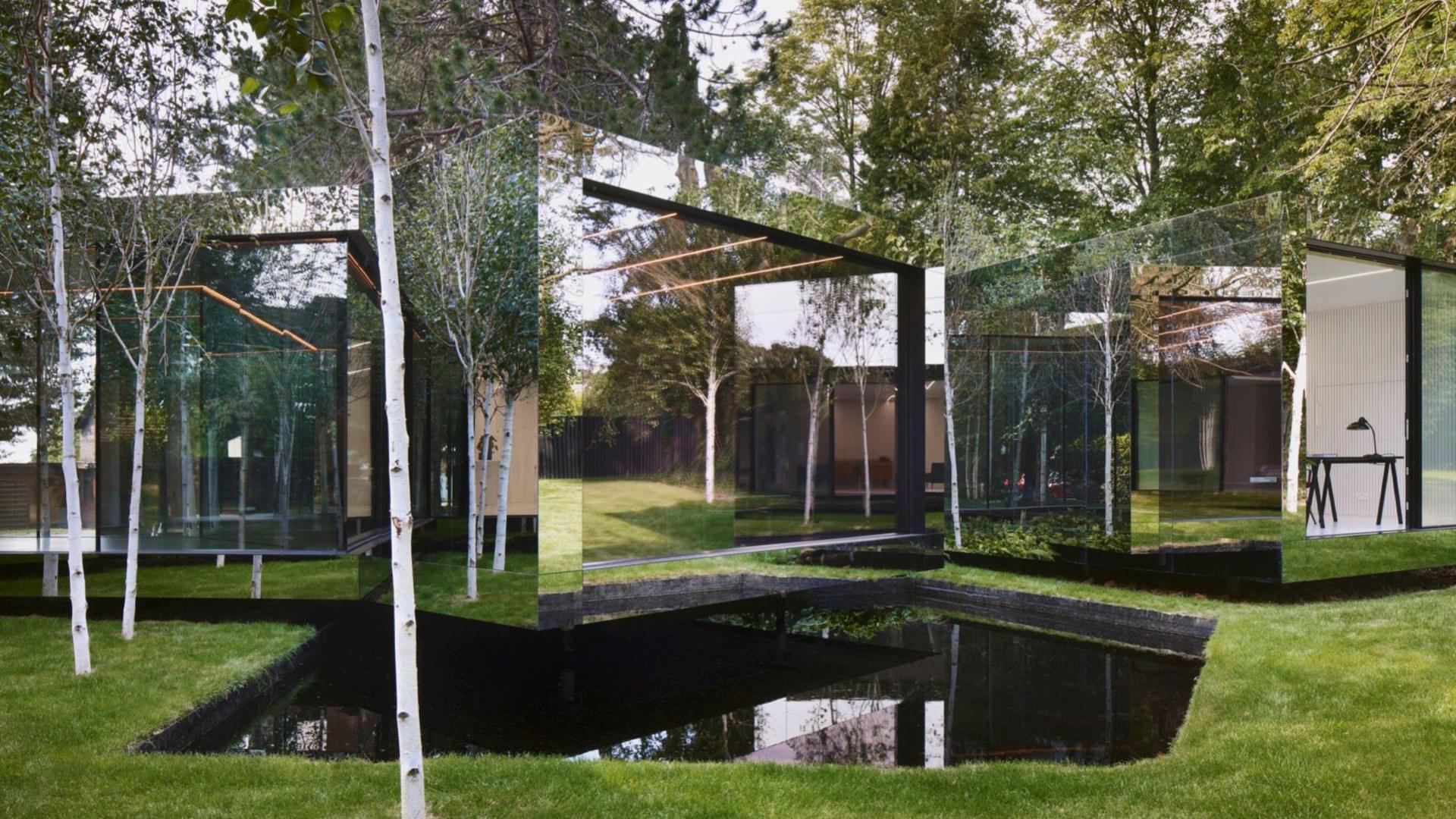 A building that is made from a mirrored material. Part of the one-storey building sits on a patch of grass and another part is elevated over a small pond. The house is surrounded by trees.