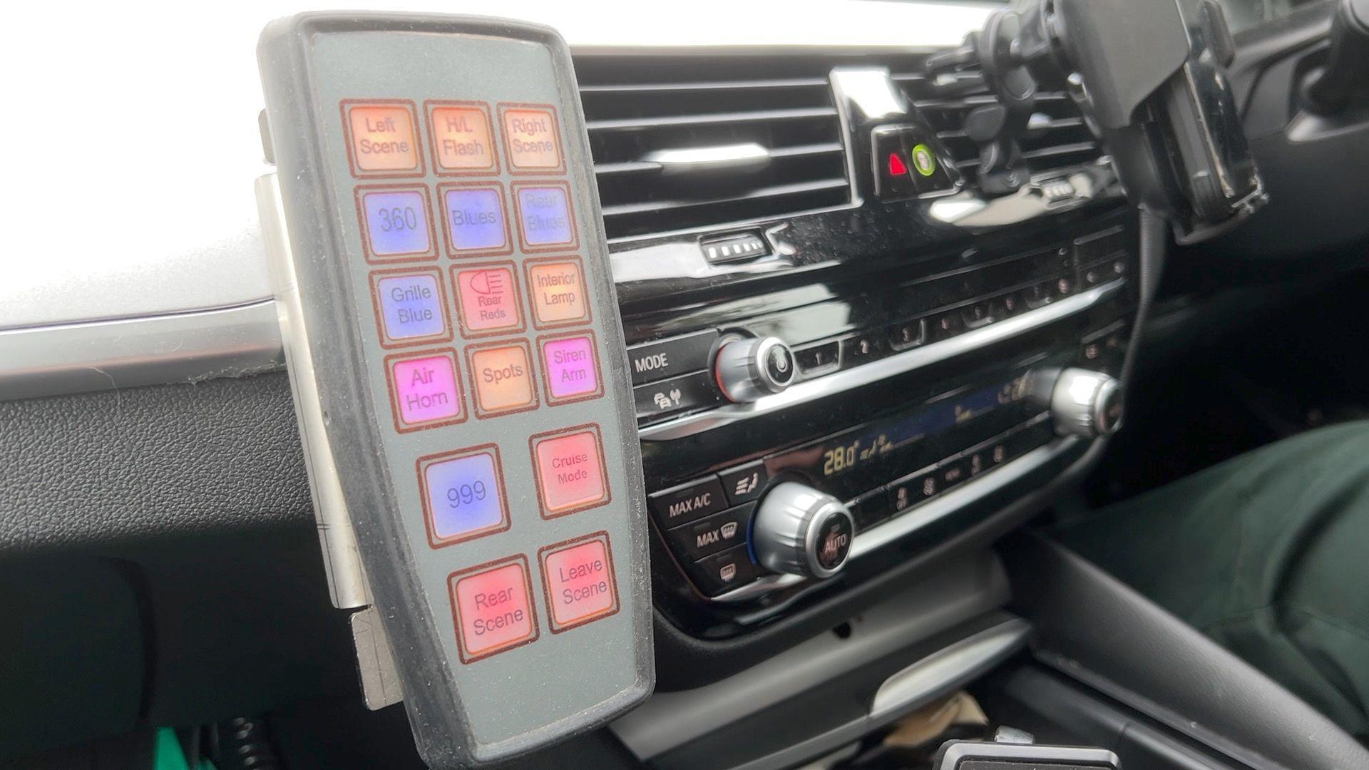 A control panel attached to the dashboard of a car, with buttons in bright colours - orange, blue, pink, and red. The buttons all say different thinks on them - one says Blues and one says 999.