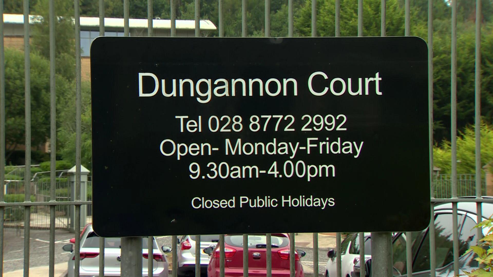 A black sign saying 'Dungannon Court' with the opening hours on it hangs on a silver metal gate. Behind the gate is cars parked in a car park.