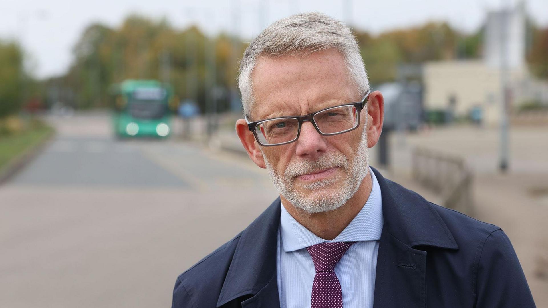 Darren Roe is looking at the camera. He has short, grey hair and a beard and moustache. He is wearing spectacles, a light-coloured shirt, burgundy tie and a blue coat. In the background is a blurred image of a bus.