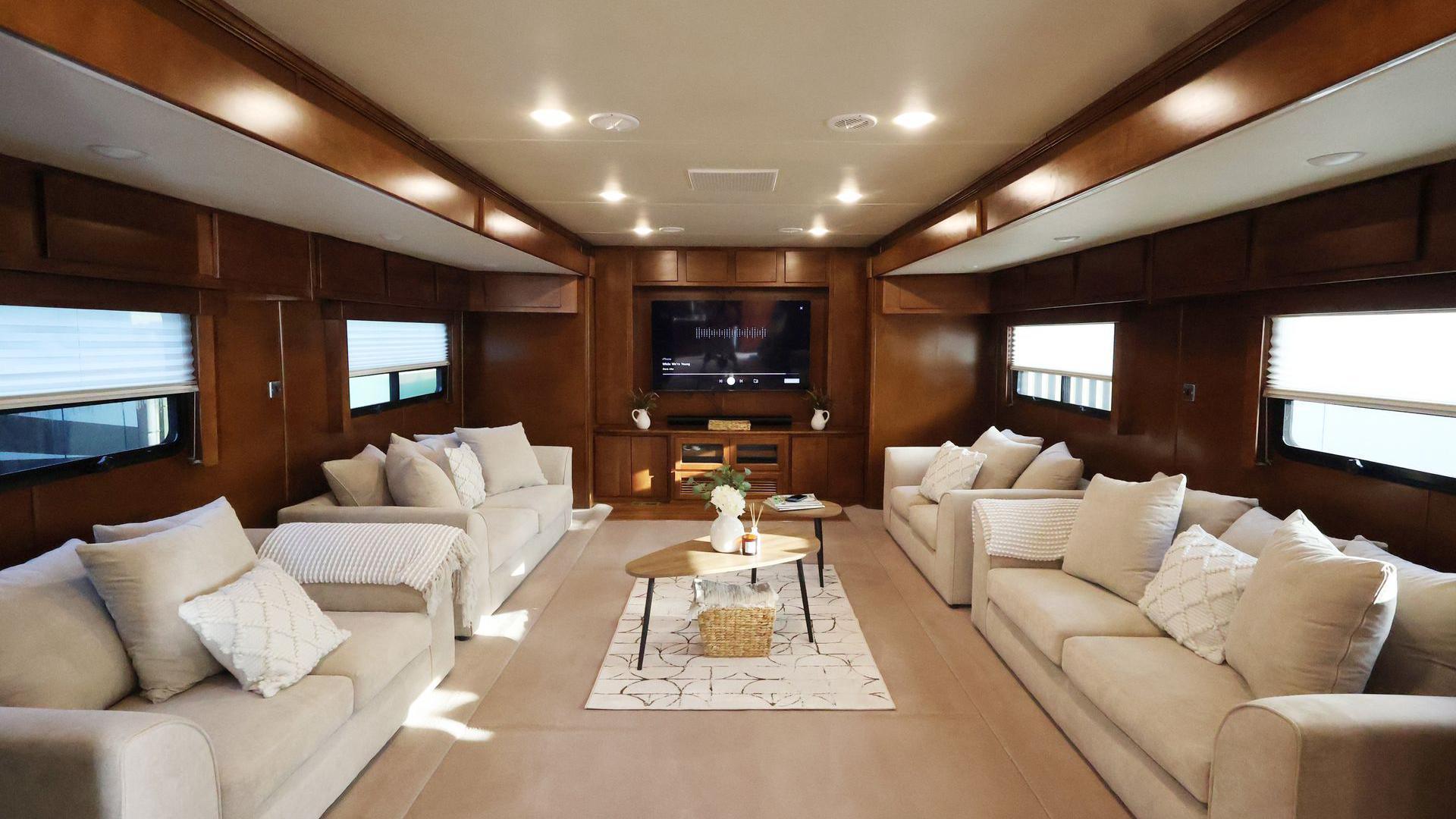 Four large white sofas with pillows inside a custom-made trailer. In the middle of the room are two coffee tables with a vase on top. There is also a wide screen television mounted on the wall. 