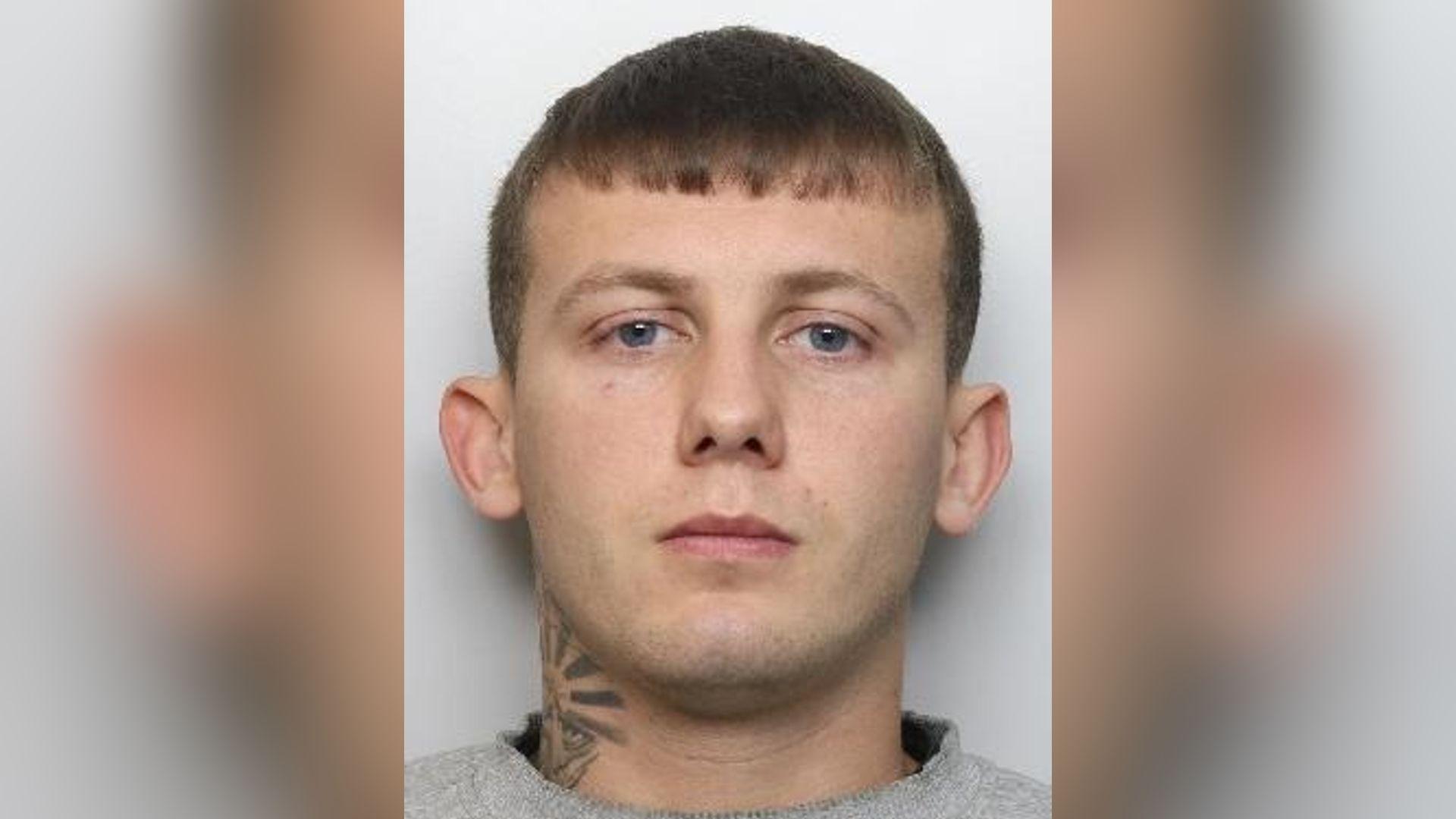 A mugshot of a white man with neck tattoos looks towards the camera. He is wearing a grey jumper.