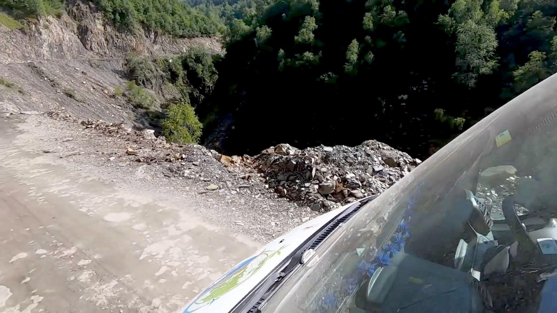 A view taken from outside the window of a camper van showing a very steep cliff drop off a dirt road, at the bottom of the drop is a woodland. The windscreen of the camper van is visible in the right of the image