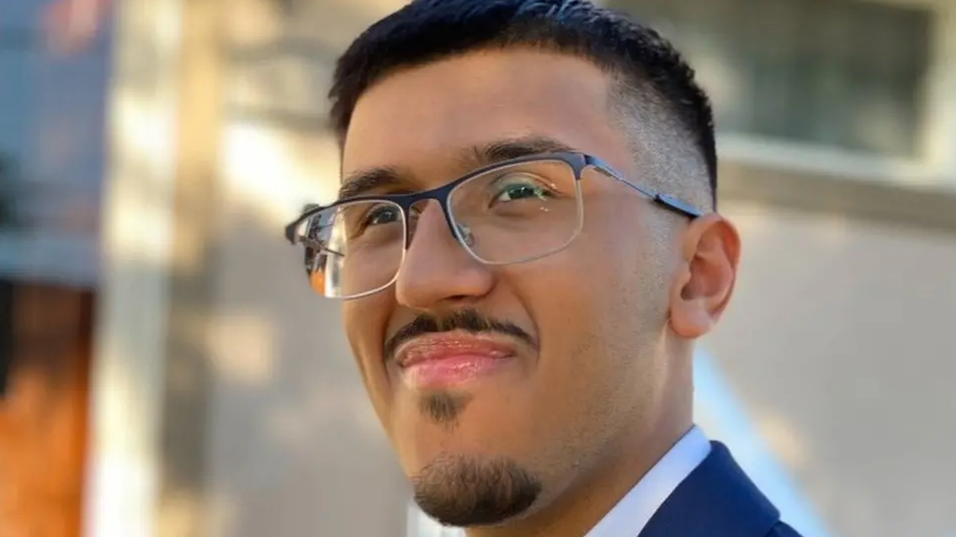 A head and shoulders photo of Sachin Hawkins. He has short black hair, a short goatee beard and mustache, and is smiling with closed lips. He wears half-rimmed glasses, a blue suit jacket and a white shirt.