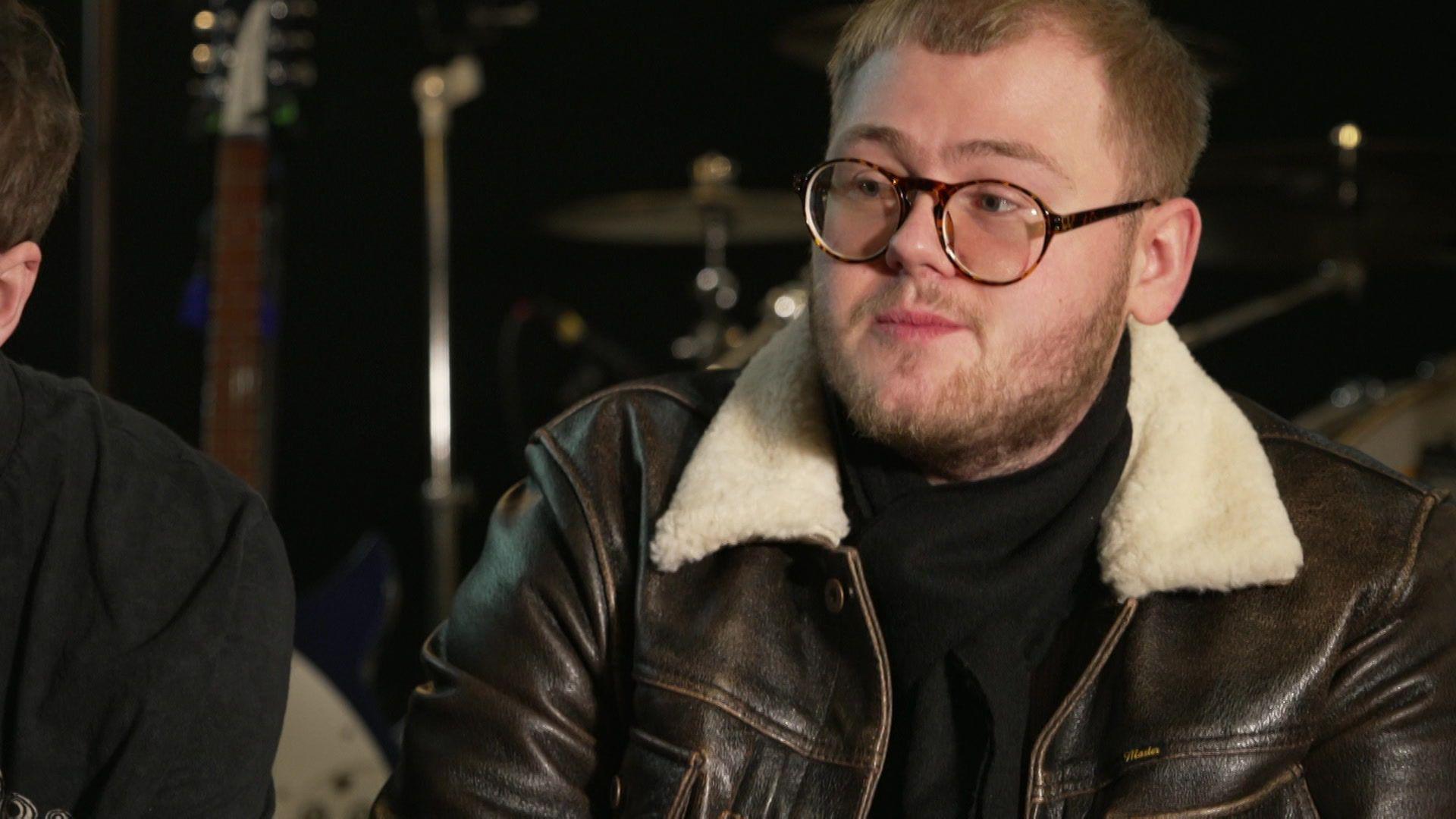 Alex Moore wearing a brown jack and brown-rimmed glasses sits in a recording studio for an interview