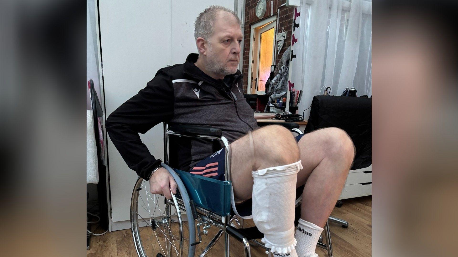 Rob Taylor is sitting in a wheelchair, he wears navy shorts with pink stripes and a black zipped jumper. He has a bandage on his right leg and is holding the wheel of his chair.