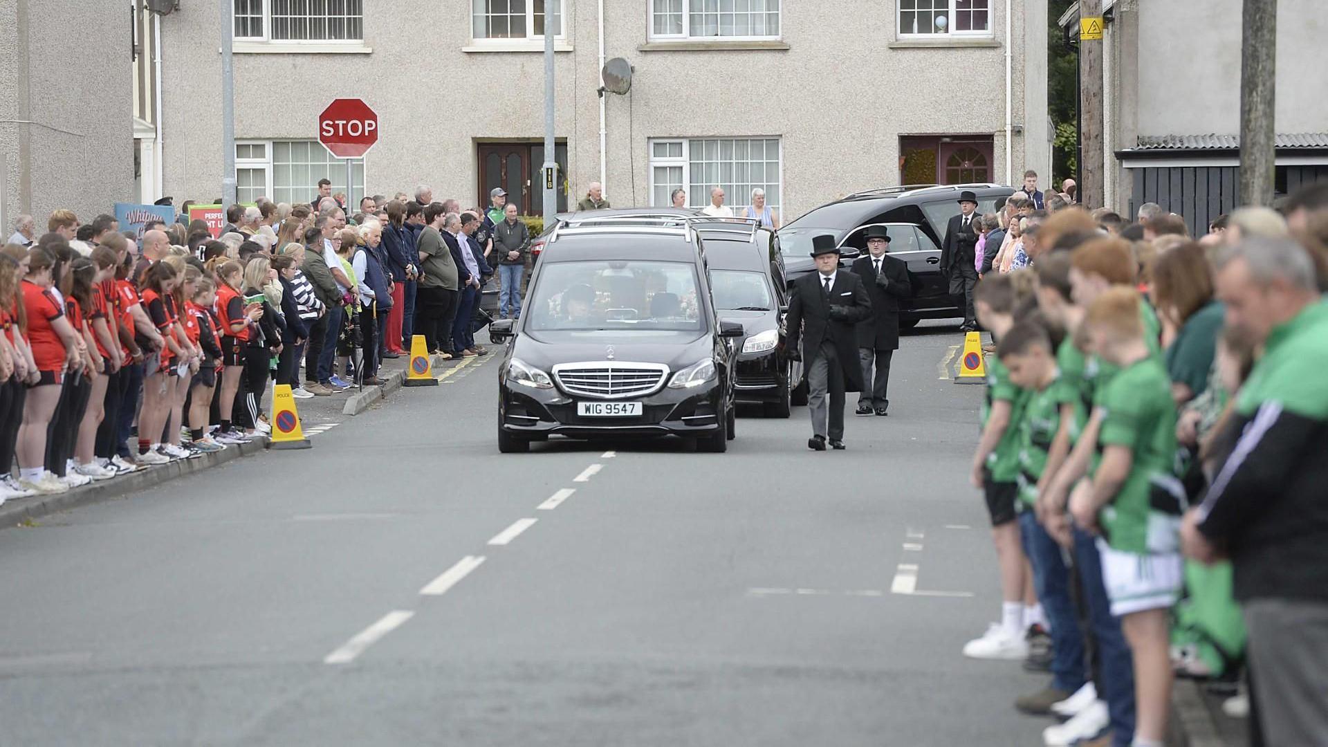 Three hearses drive down a road. On either side, large crowds of people watch. 
