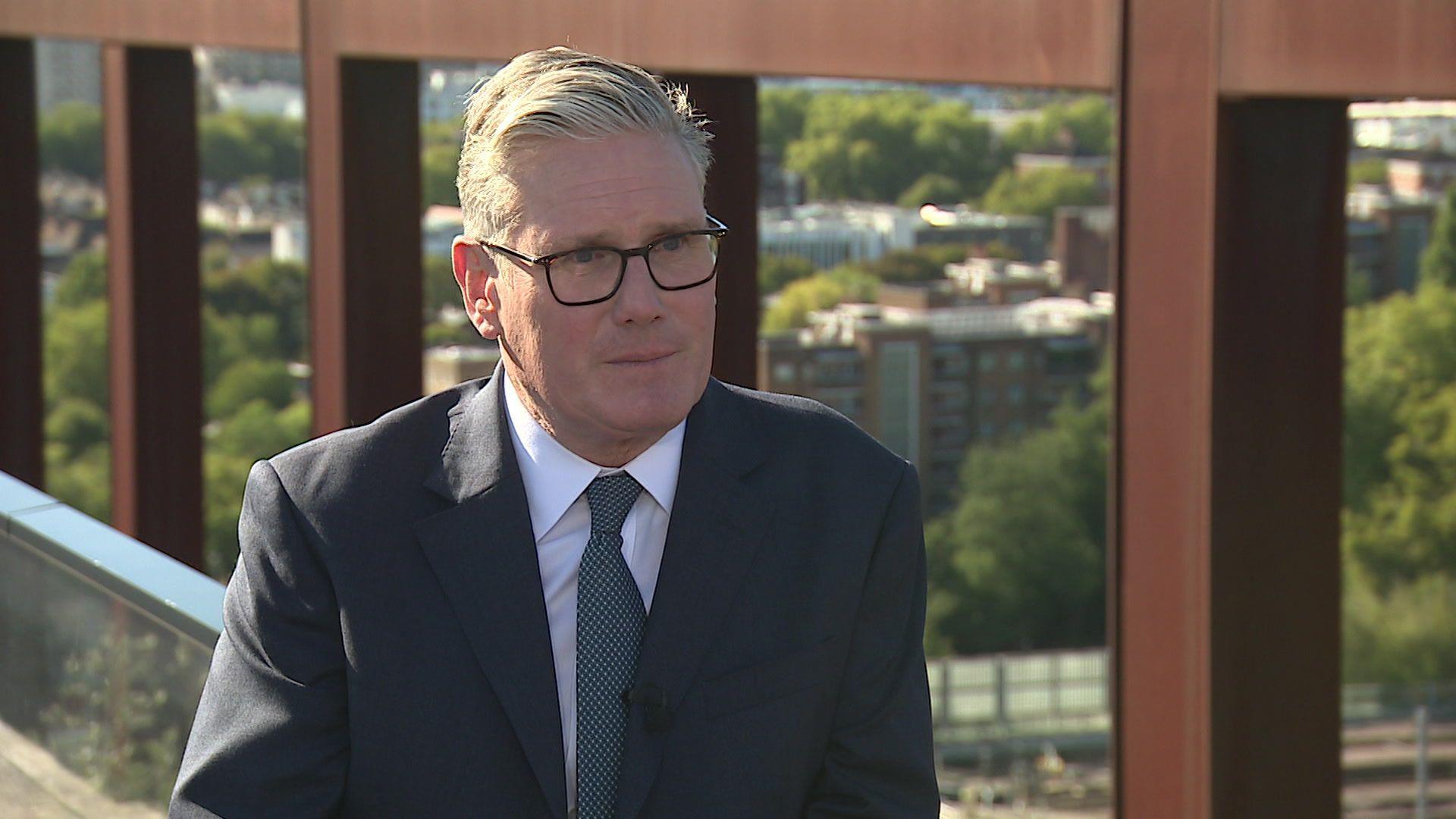 Sir Keir Starmer sat by a balcony overlooking part of London, with a bridge in the background. He is wearing a blue suit and blue tie.