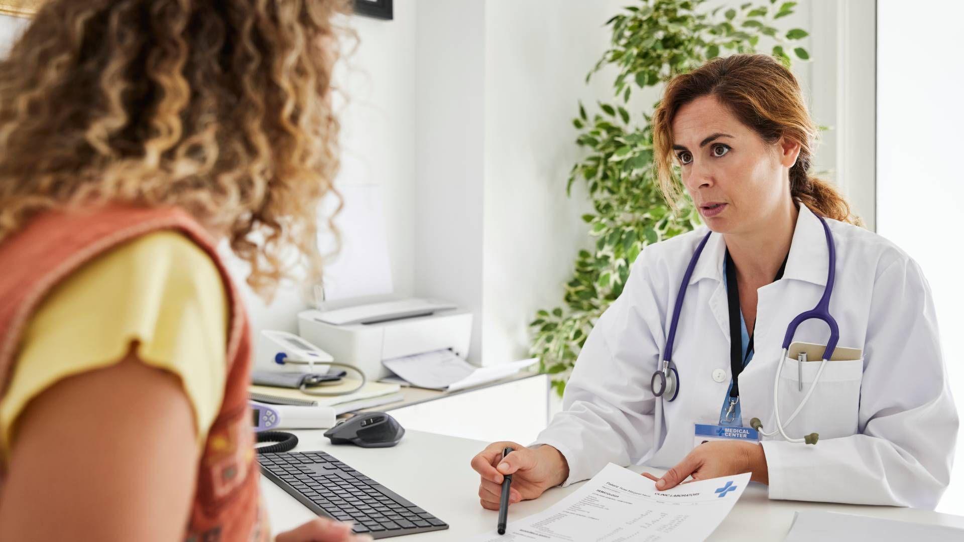 A female doctor writes on a piece of paper and looks at her patient, a woman with curly blonde hair. The doctor is pointing at a piece of paper on the desk with a pen.