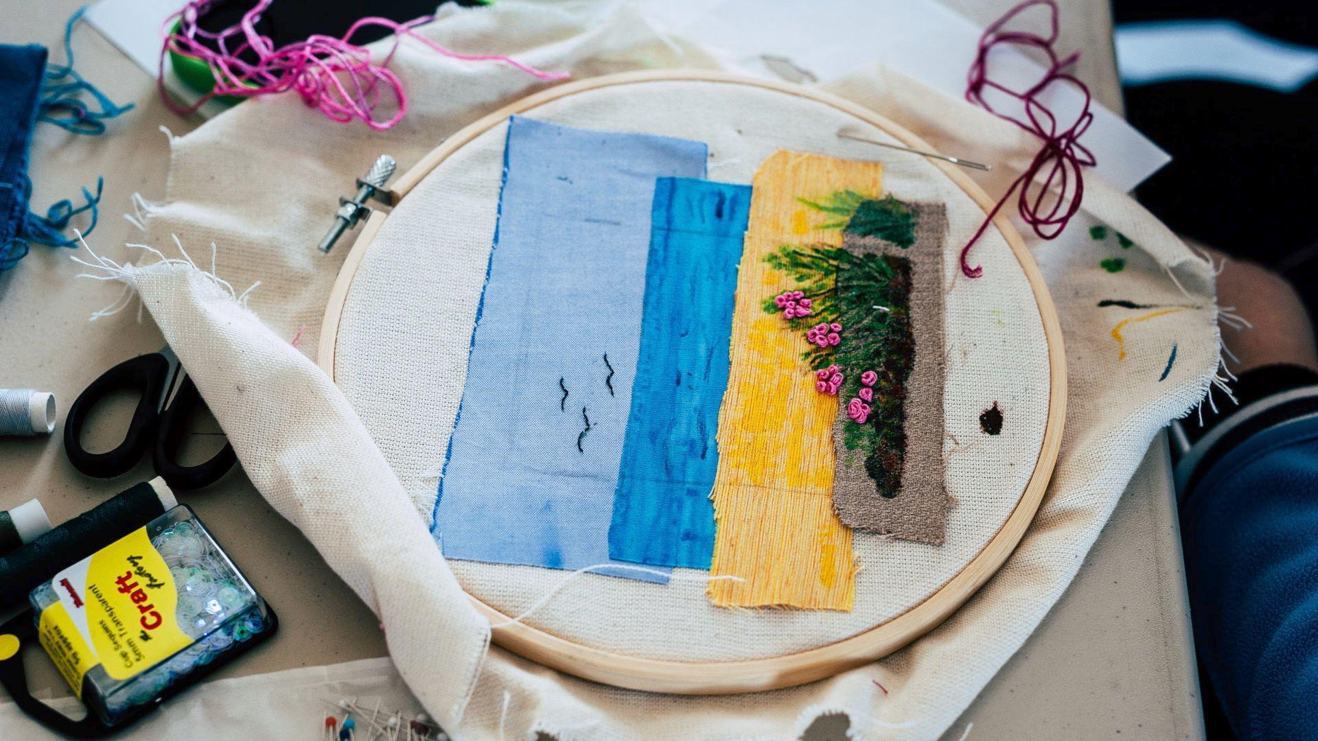 A close up of a piece of embroidery, still in the wooden frame, surrounded by sewing materials like scissors and thread. The embroidery depicts a beach scene with blue sky, birds flying, yellow sand and some pink flowers.