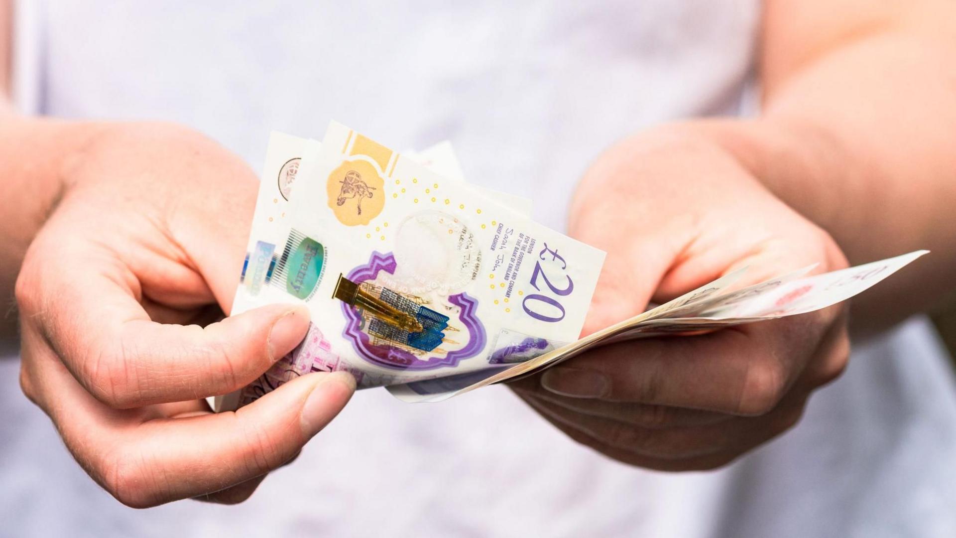 A woman counting UK sterling bank notes in her hands. At least one of them is a purple £20 note.