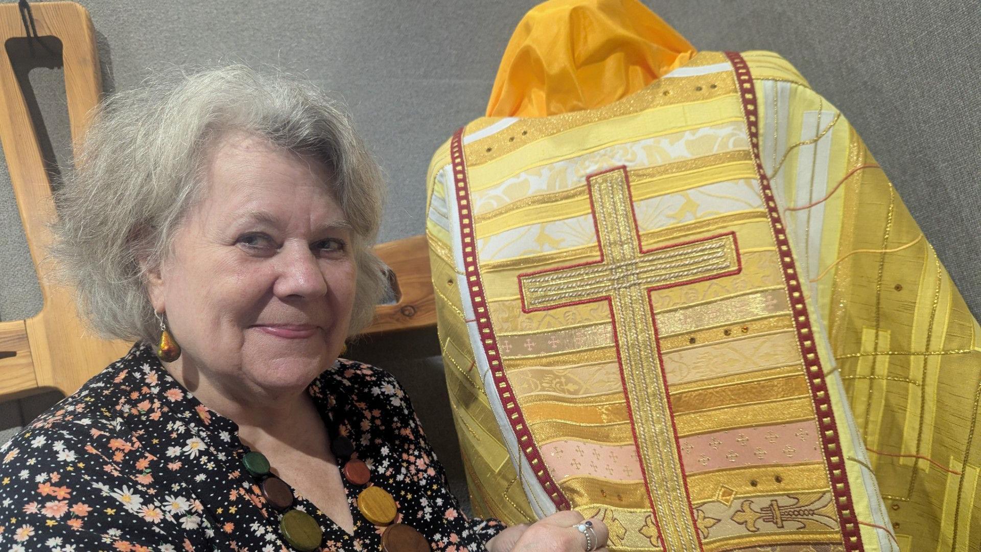 Juliet Hemingway with grey hair wearing a flower-patterned dress and holding a yellow clerical cloak with a large cross embroidered onto it.