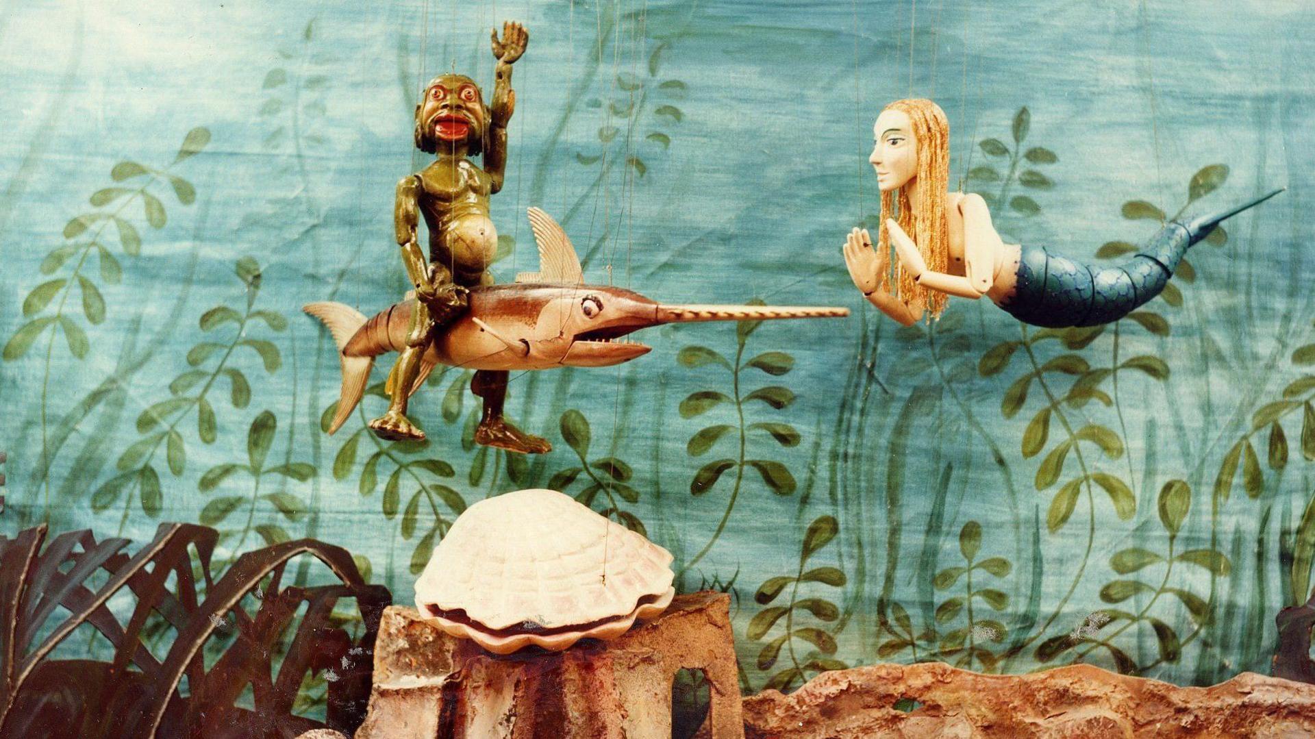 A colour photograph of puppets in an underwater scene. There is a mermaid and a green humanoid creature riding a swordfish.