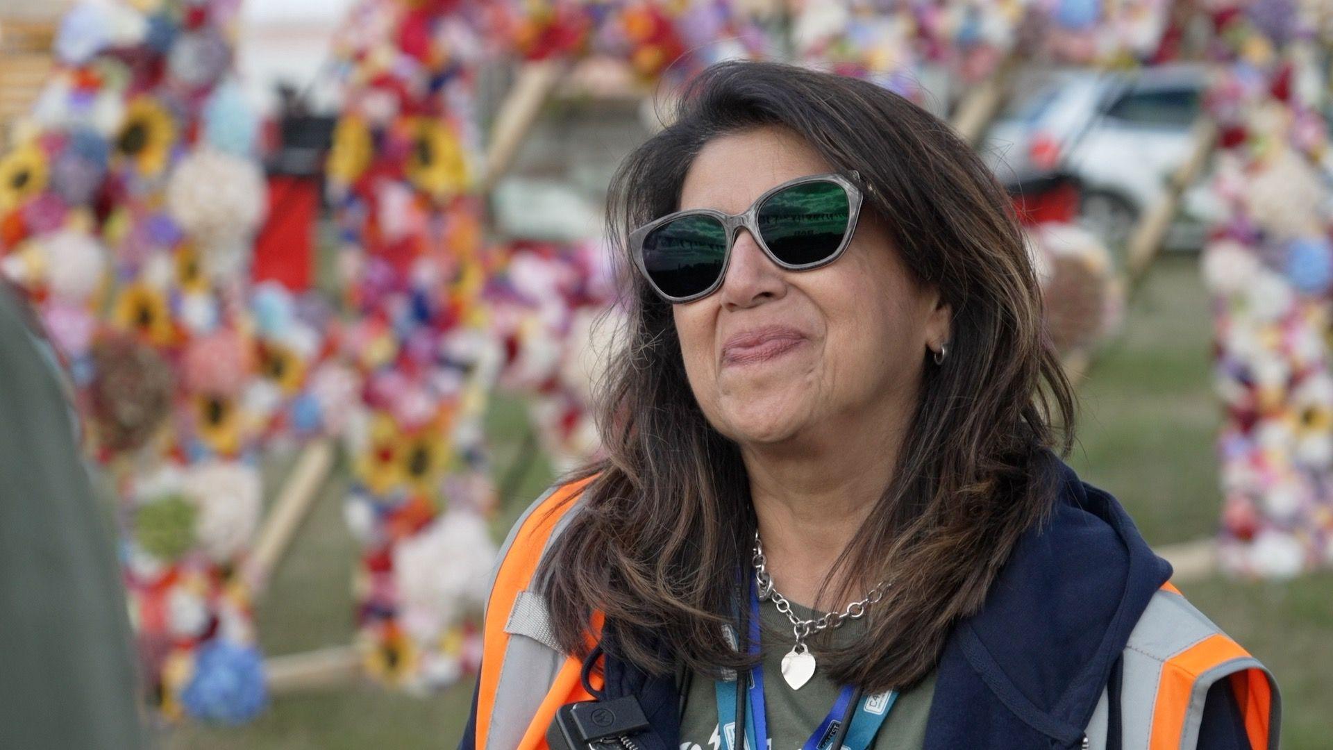 A woman with brown hair is wearing sunglasses and smiling. There are flowers behind her and she is wearing a high vis.