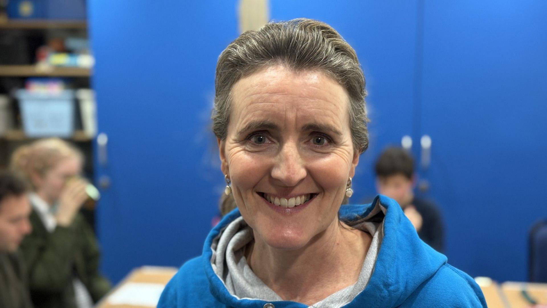 A woman wearing a blue hoodie smiles at the camera. She has dark hair that has been tied back. 