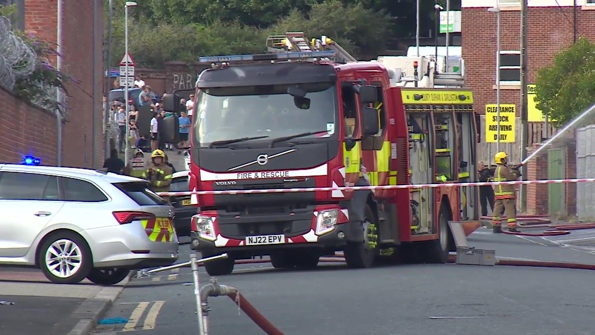 A fire truck sits behind a cordon. Crowds of people can be seen on the left and a firefighter pointing a hose at a building can be seen on the right.